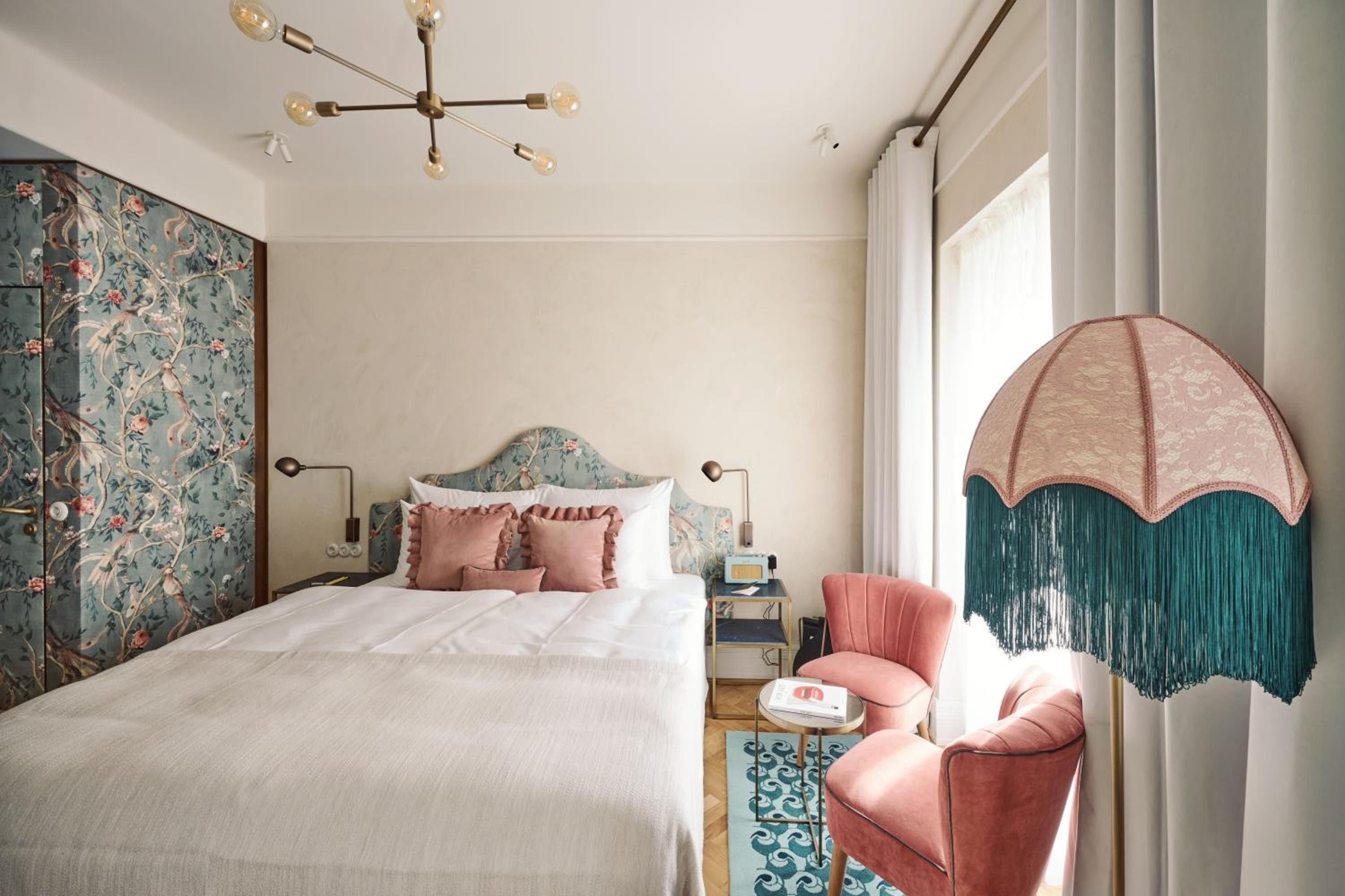 Whimsical chinoiserie wallpaper meets mid-century brass fixtures in this playfully sophisticated guest room, where coral velvet seating and a fringed parasol create intimate charm.