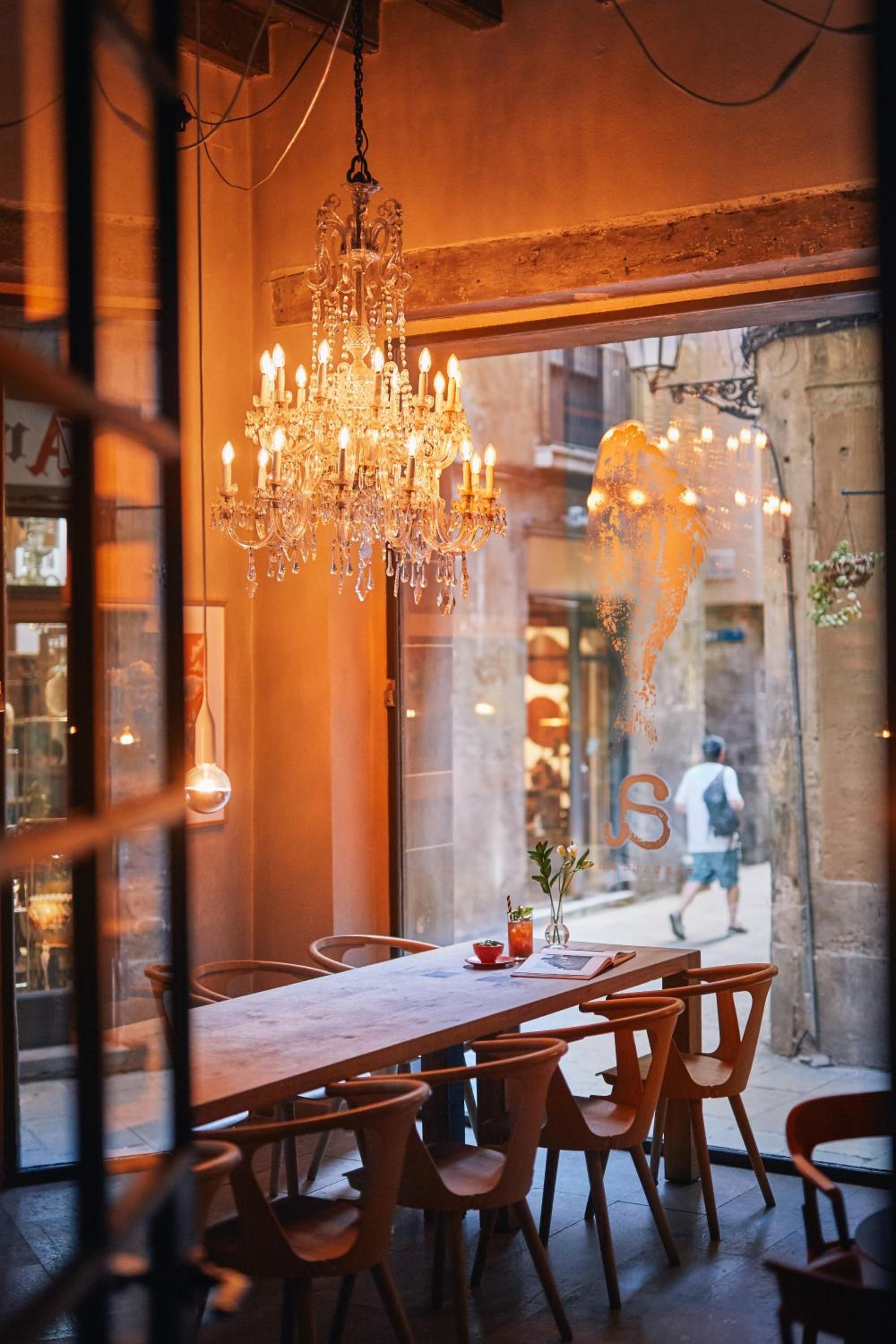 Crystal chandelier casts golden light over weathered wooden tables in this intimate dining nook, where centuries-old stone walls frame views of Barcelona's Gothic Quarter streets.