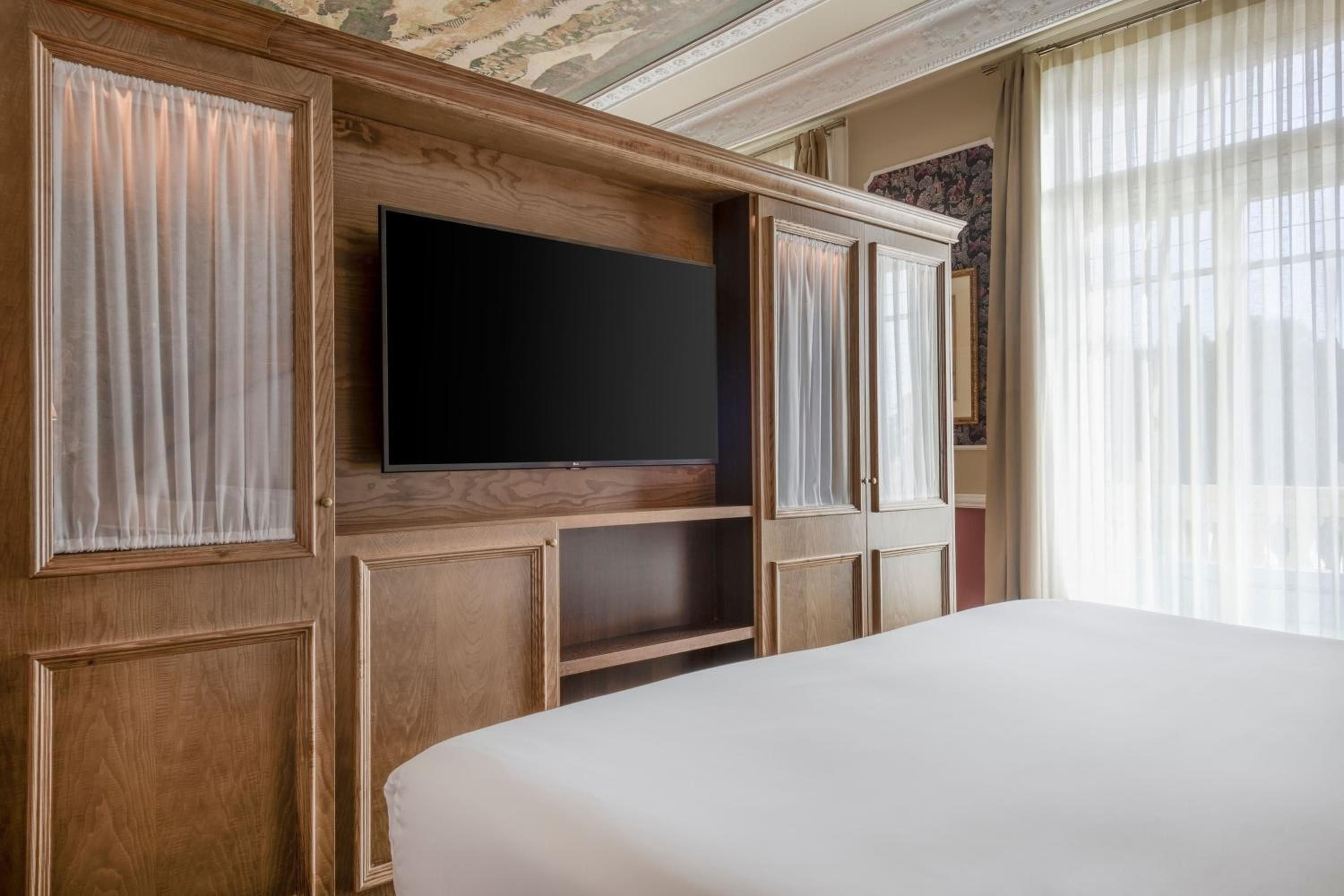 Hand-carved oak cabinetry frames a sleek television mount, while gauzy curtains filter Madrid sunlight across pristine linens in this thoughtfully appointed guest room.