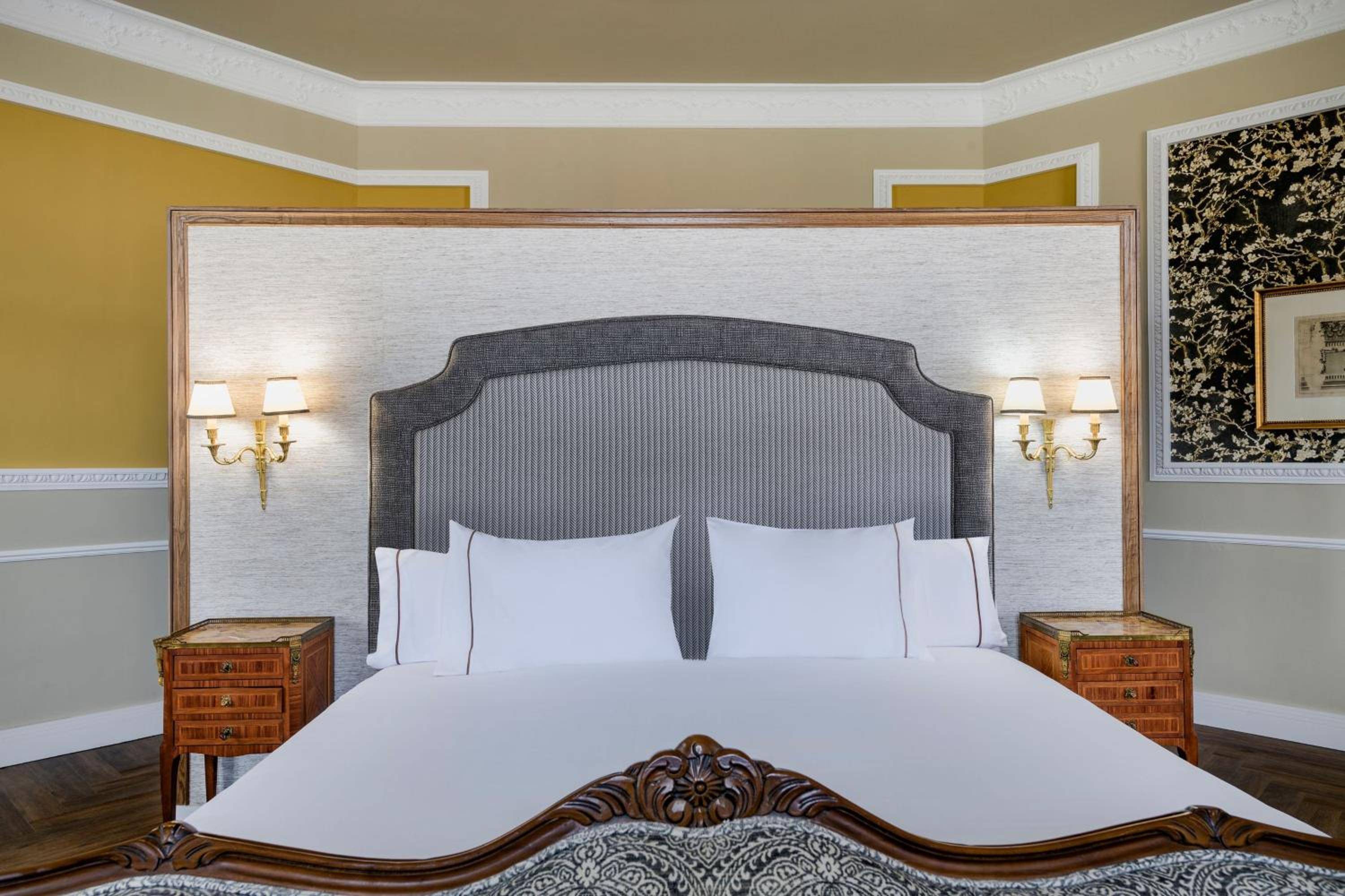 Sophisticated gray pinstripe headboard anchors this elegant guest room, where brass sconces cast warm light against textured wallpaper and rich wooden nightstands.