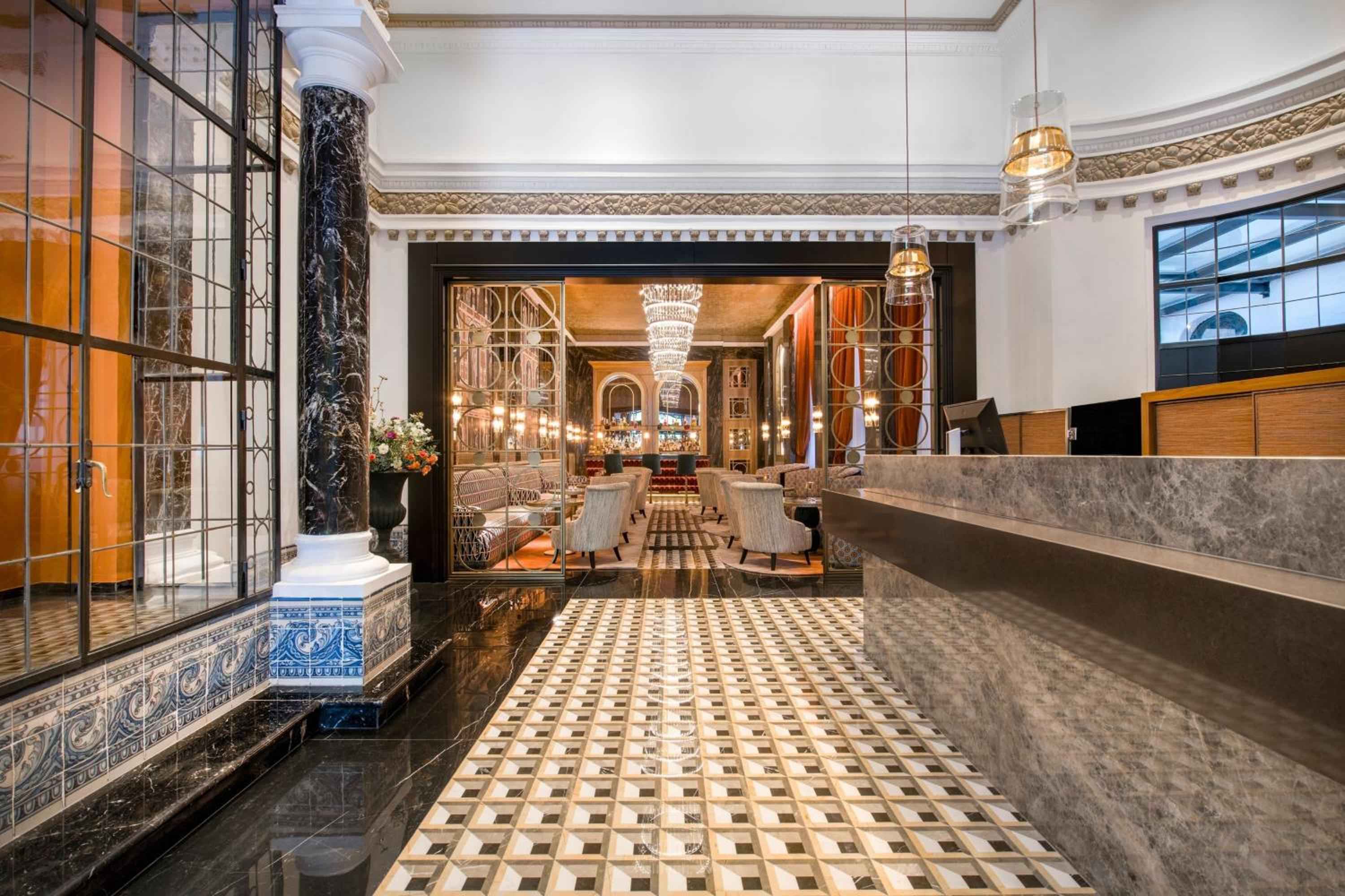 Geometric marble floors lead through a grand lobby where classical columns meet contemporary elegance, crystal chandeliers casting warm light over polished surfaces and plush seating areas.