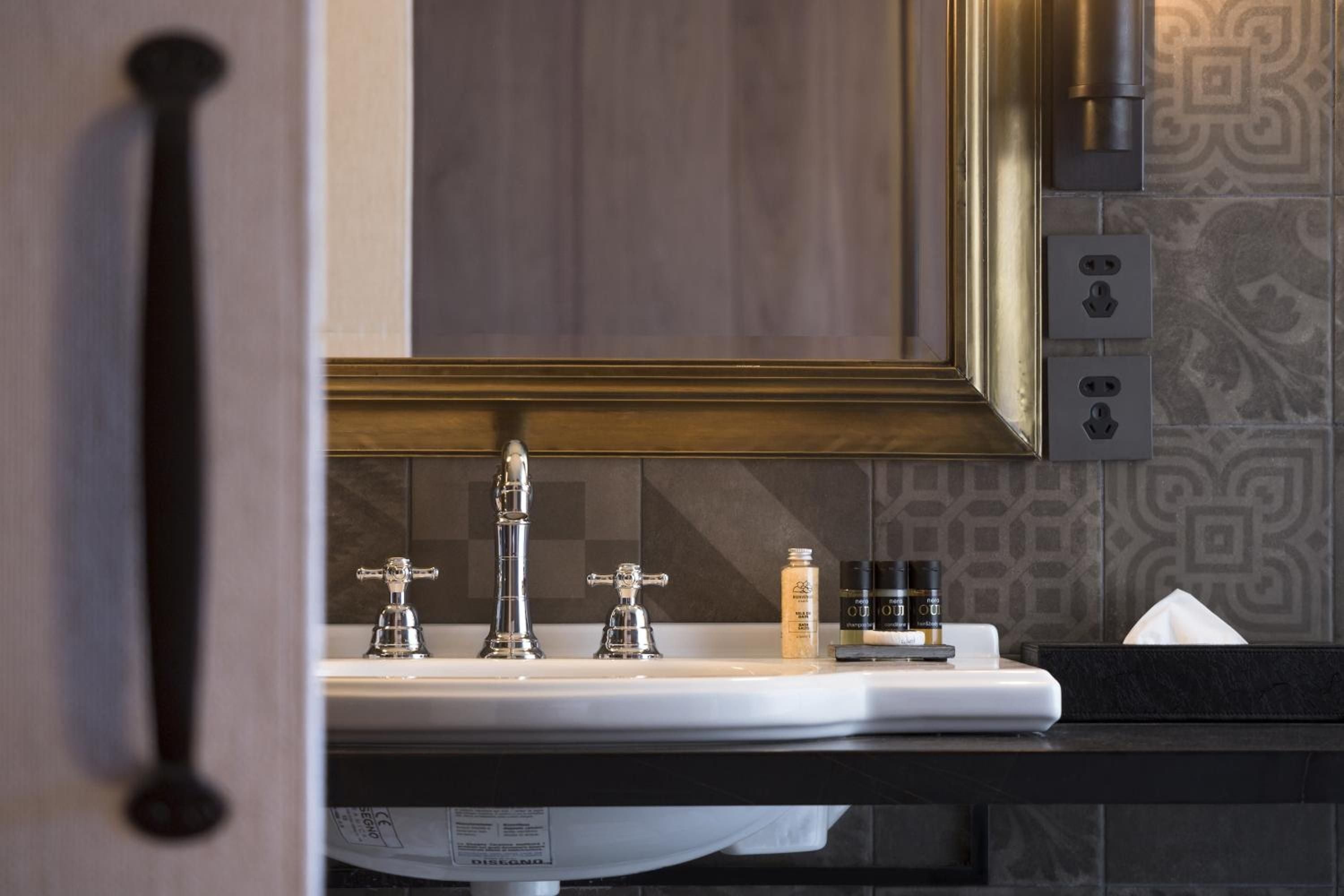 Polished chrome fixtures and geometric patterned tiles create sophisticated contrast against warm golden mirror frames, while curated amenities add thoughtful luxury touches.