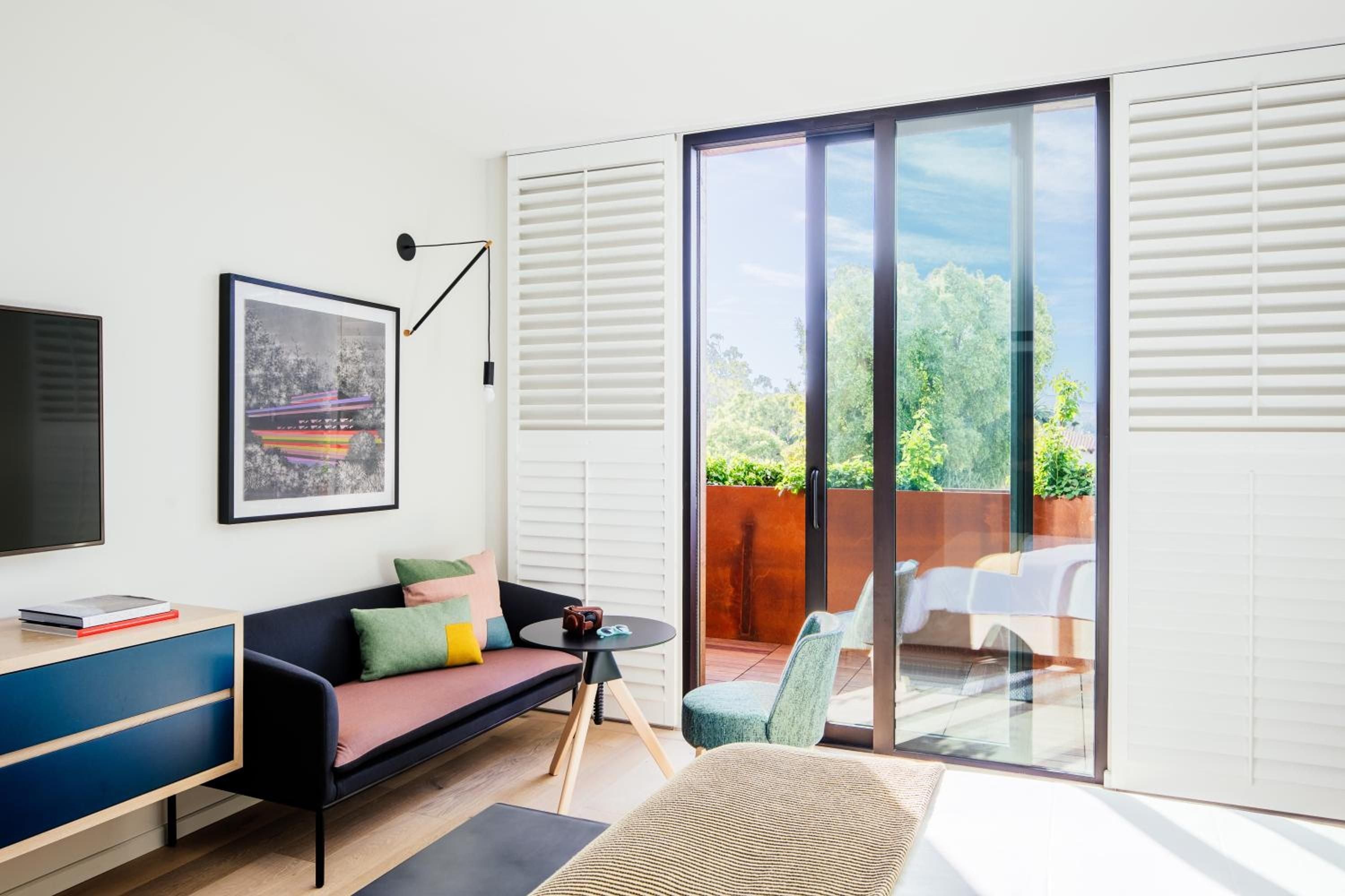 Mid-century furnishings and jewel-toned accents create a sophisticated retreat, while floor-to-ceiling glass doors frame the terrace's sun-dappled seating and verdant hillside views.