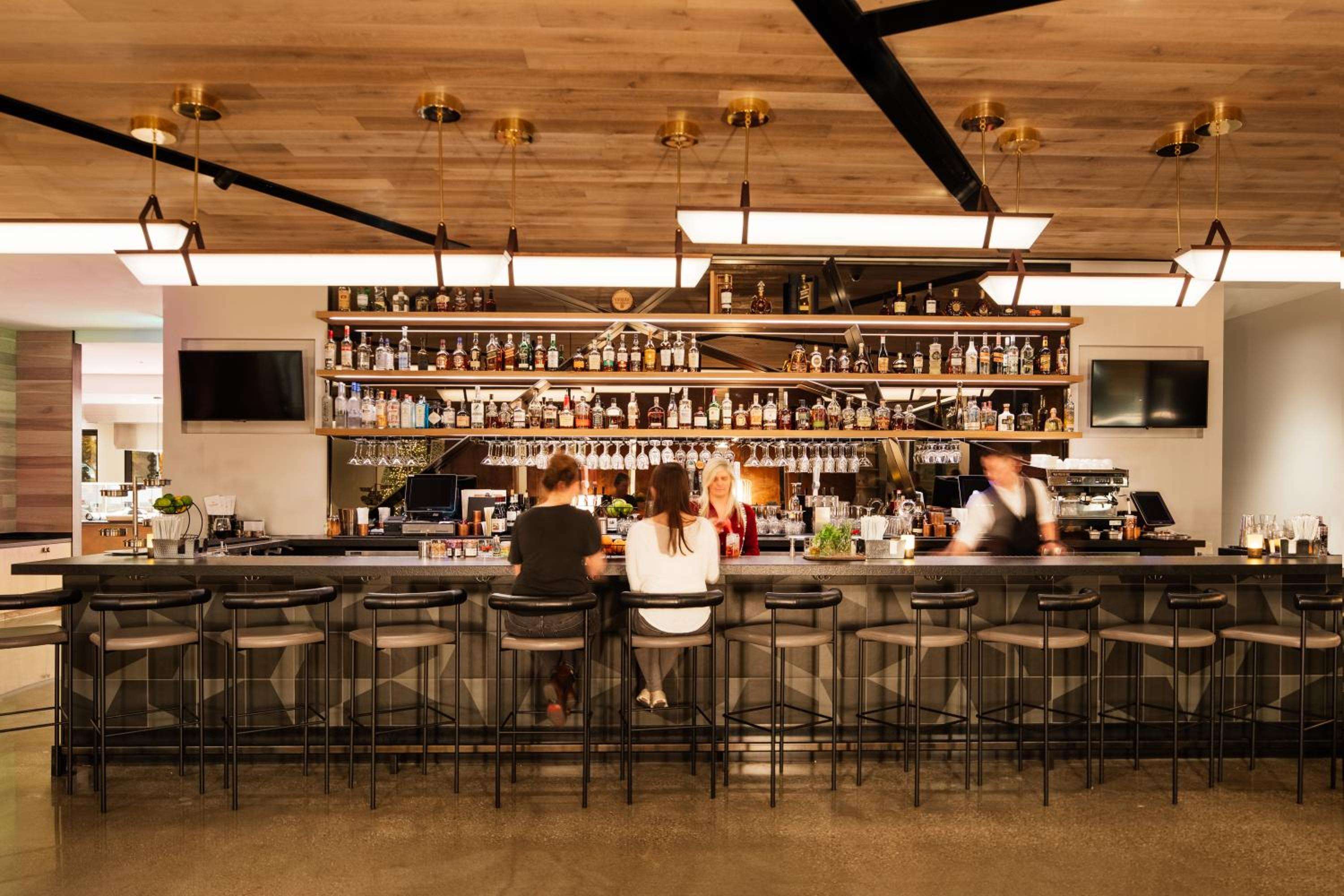 Warm pendant lights cast a golden glow over the sleek marble bar, where craft cocktails flow beneath shelves lined with premium spirits and hanging glassware.