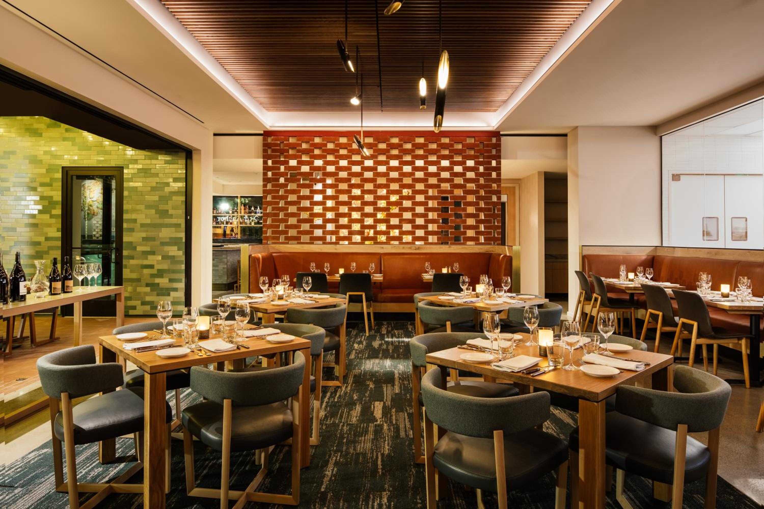 Warm wood slats and copper-toned tiles create intimate dining alcoves, where sage-green chairs gather around candlelit tables beneath sculptural pendant lights.