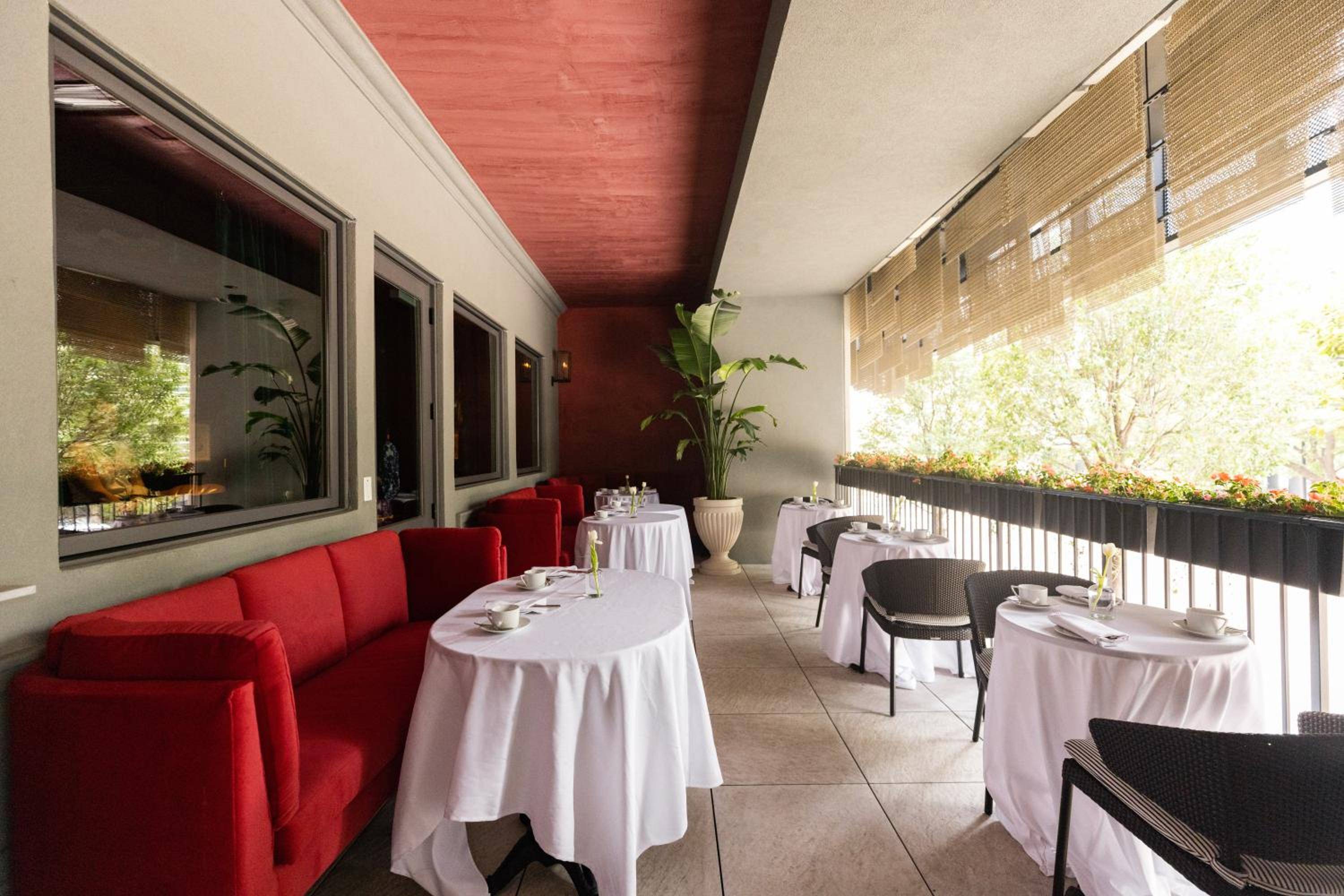 Red banquettes and crisp white linens create an intimate terrace dining experience, where bamboo screens filter dappled sunlight across thoughtfully set tables.