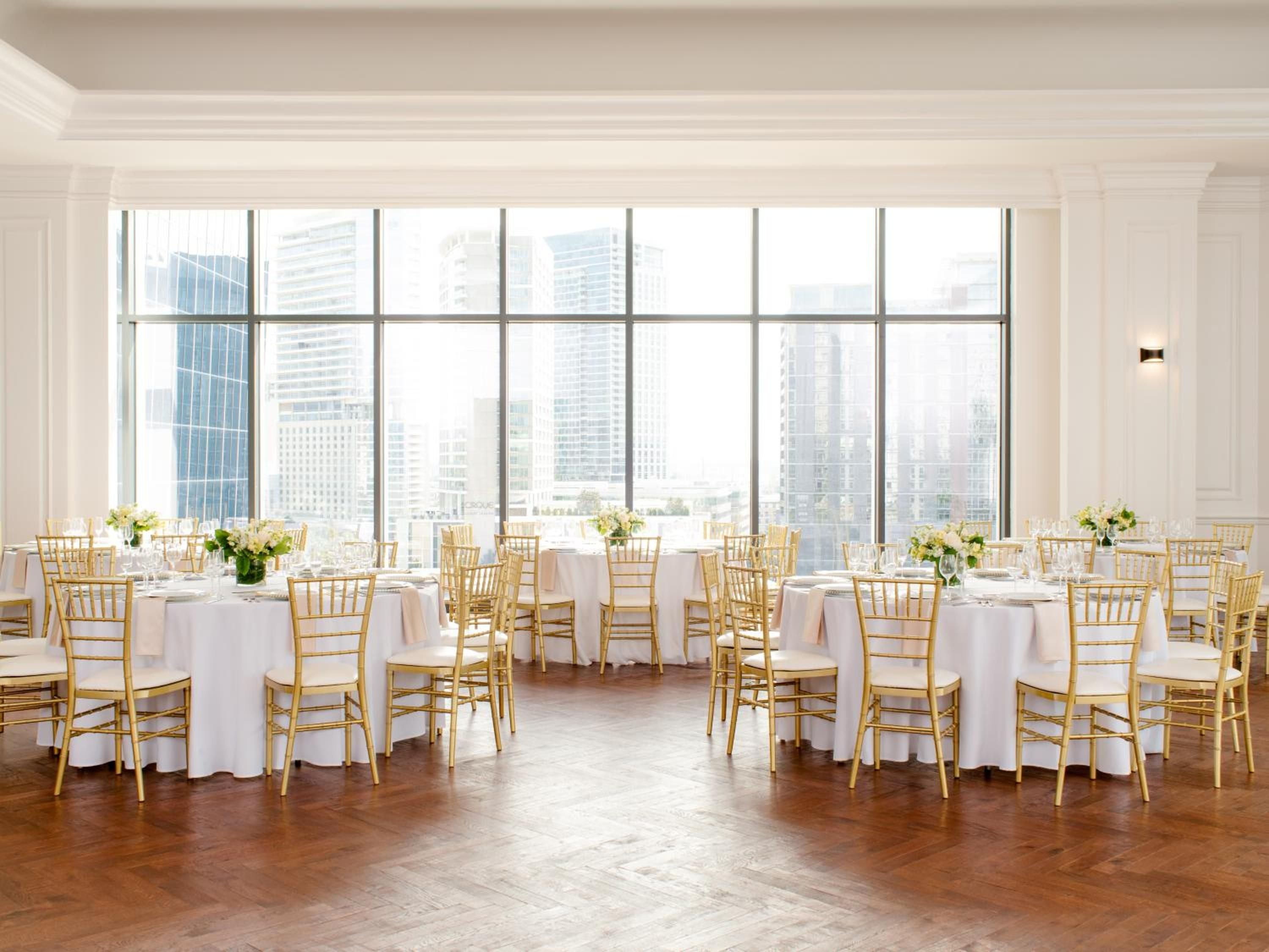 Golden Chiavari chairs and crisp white linens frame downtown Dallas through floor-to-ceiling windows, creating an elegant backdrop for celebrations high above the city.