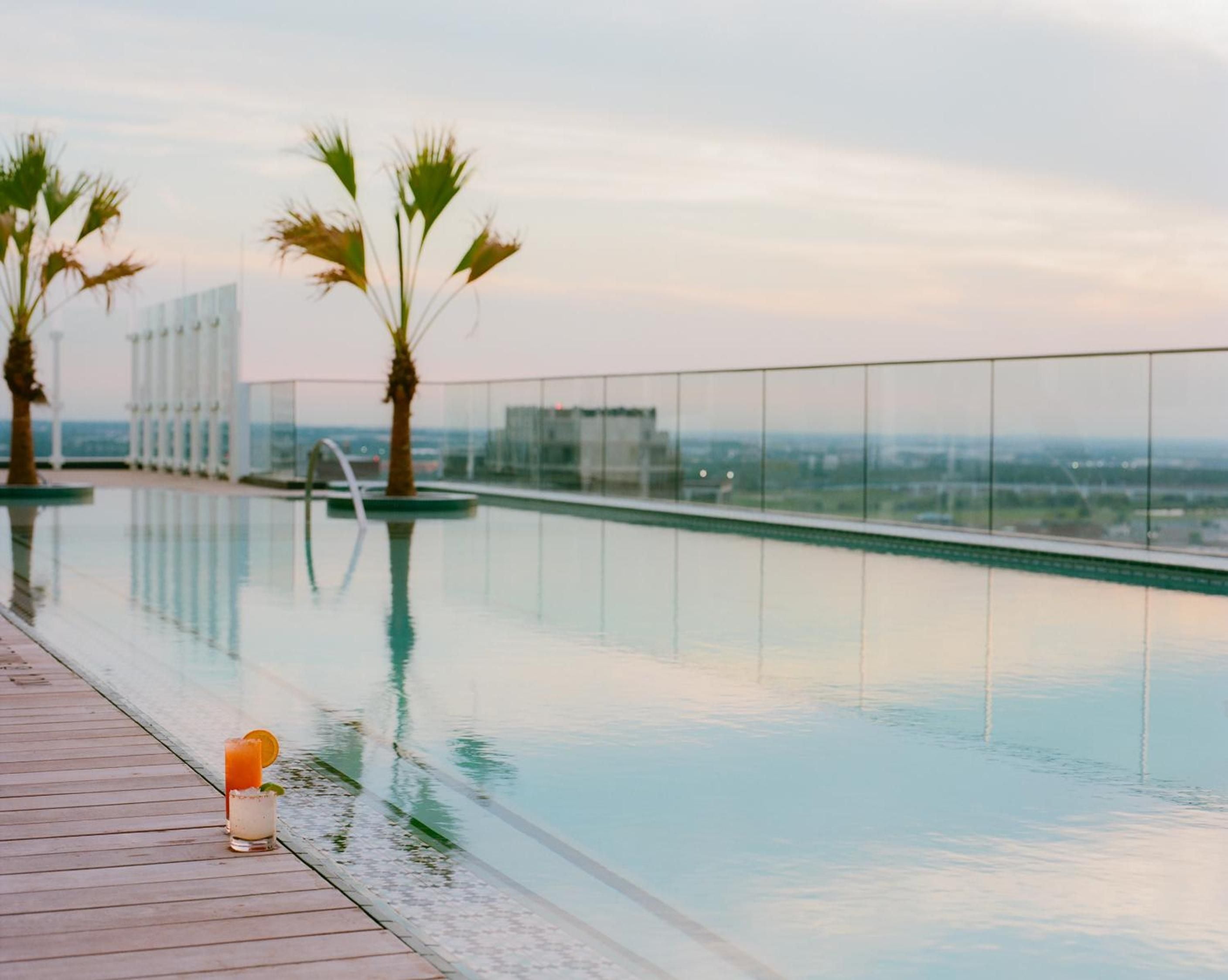 Palm trees sway beside the infinity pool as golden hour light bathes the glass-walled deck, with downtown Dallas stretching endlessly beyond.