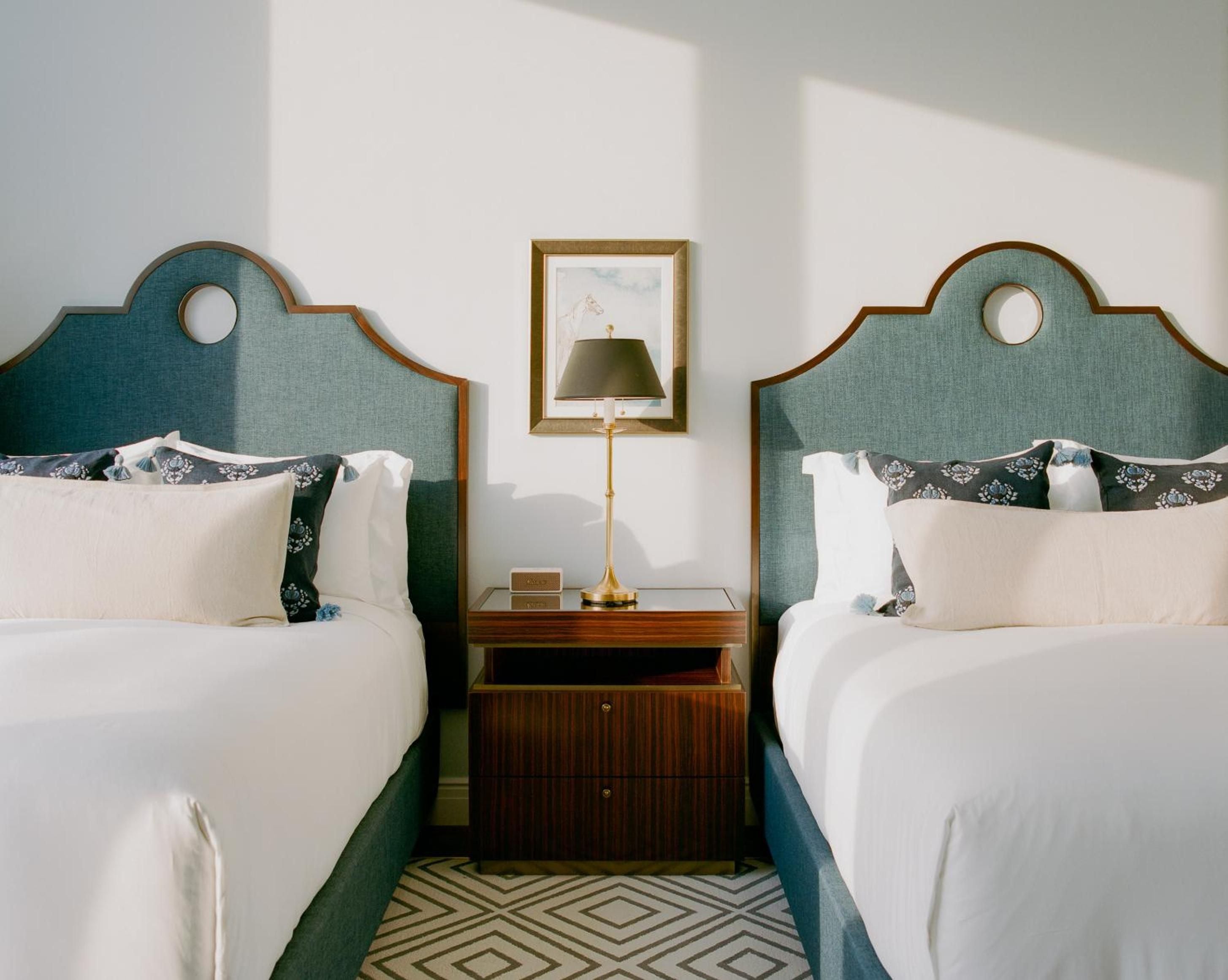 Twin beds frame a sophisticated retreat where teal upholstered headboards and geometric carpeting create perfect symmetry beneath golden afternoon light.