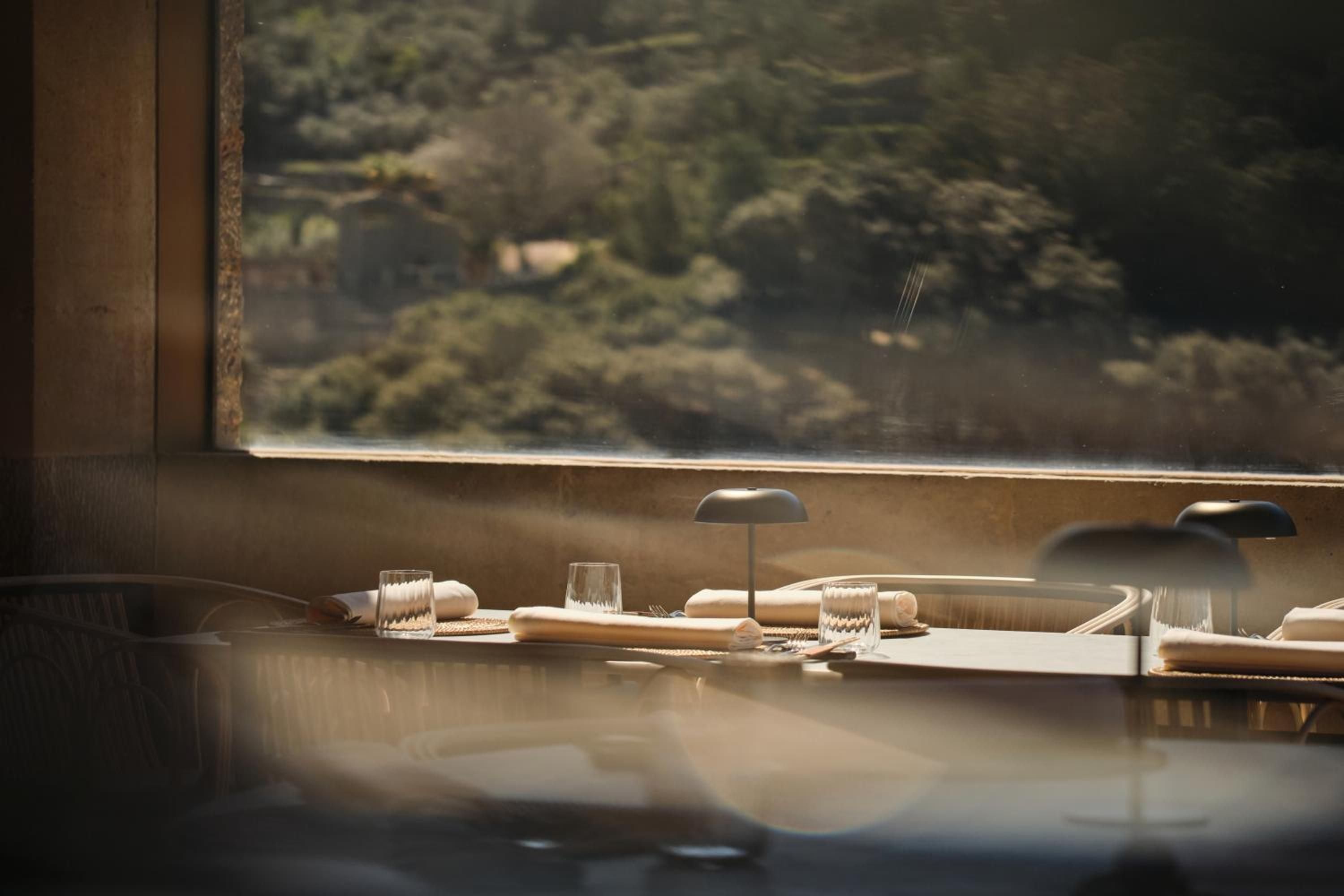 Warm afternoon light filters through expansive windows onto intimate dining tables, where crystalline glasses catch golden rays overlooking Mallorca's verdant hillside landscape.
