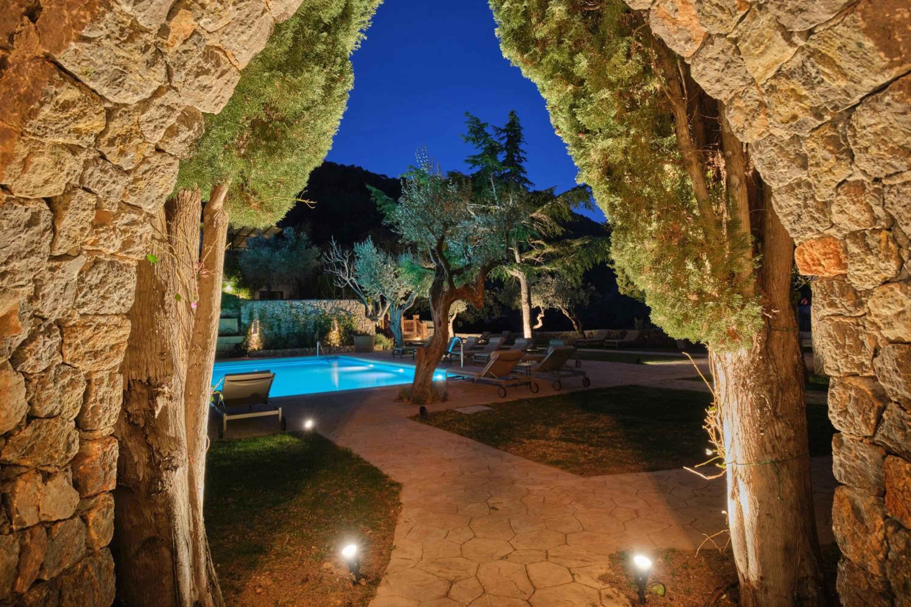 Stone archways frame an illuminated swimming pool where ancient Mallorcan architecture meets modern luxury beneath a twilight sky dotted with olive trees.