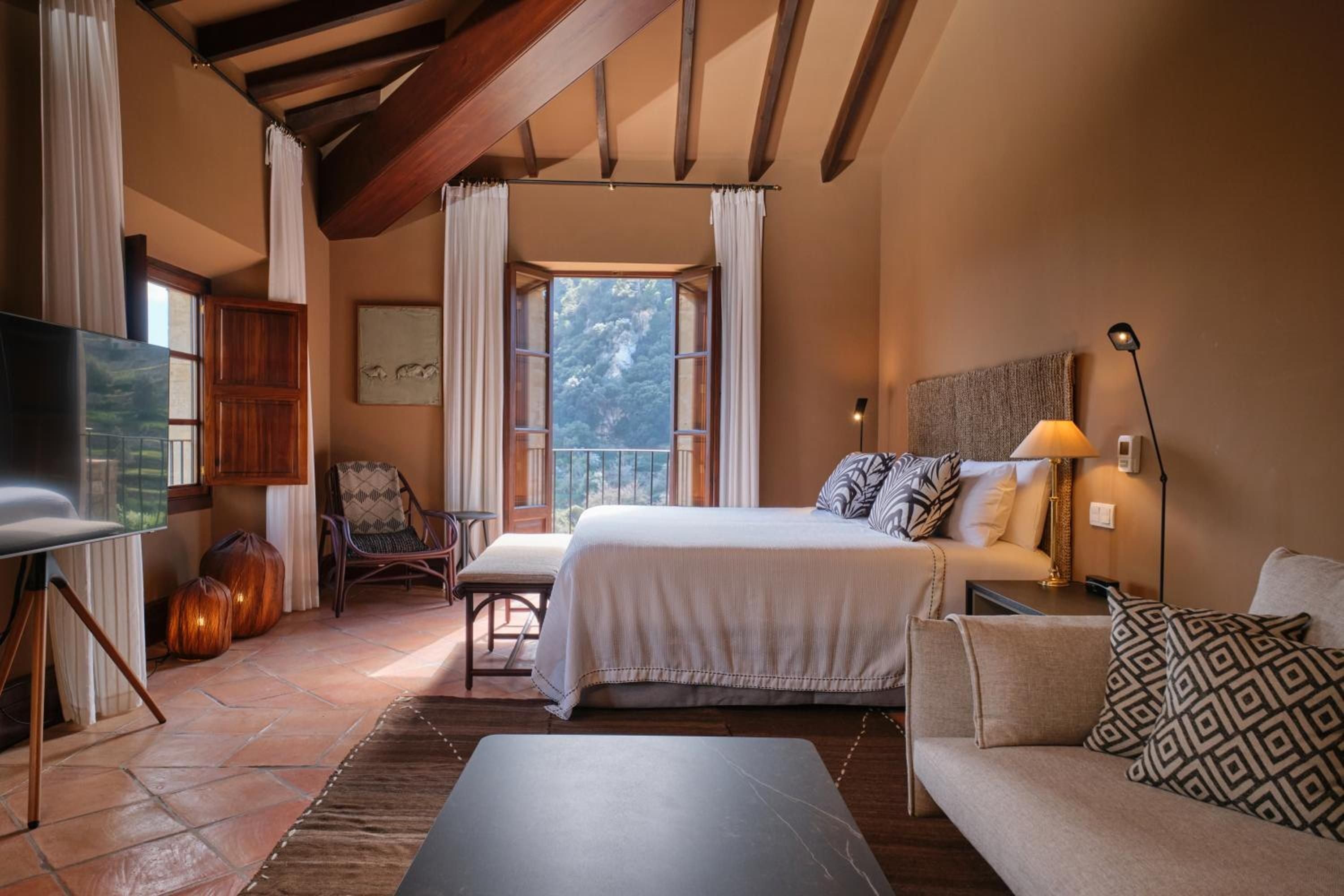 Exposed wooden beams frame this serene suite where rustic charm meets refined comfort, with French doors opening to mountain views and terracotta tiles underfoot.