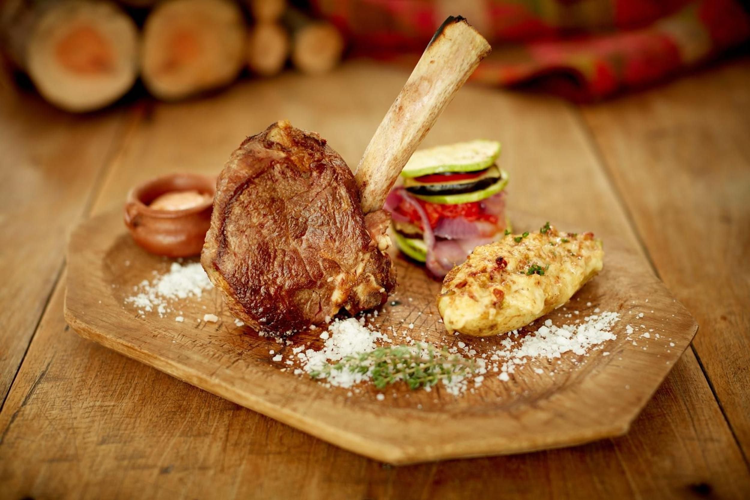 Perfectly charred tomahawk steak arrives with grilled vegetables and herb-crusted potatoes, artfully plated on rustic wood with coarse sea salt scattered like culinary confetti.