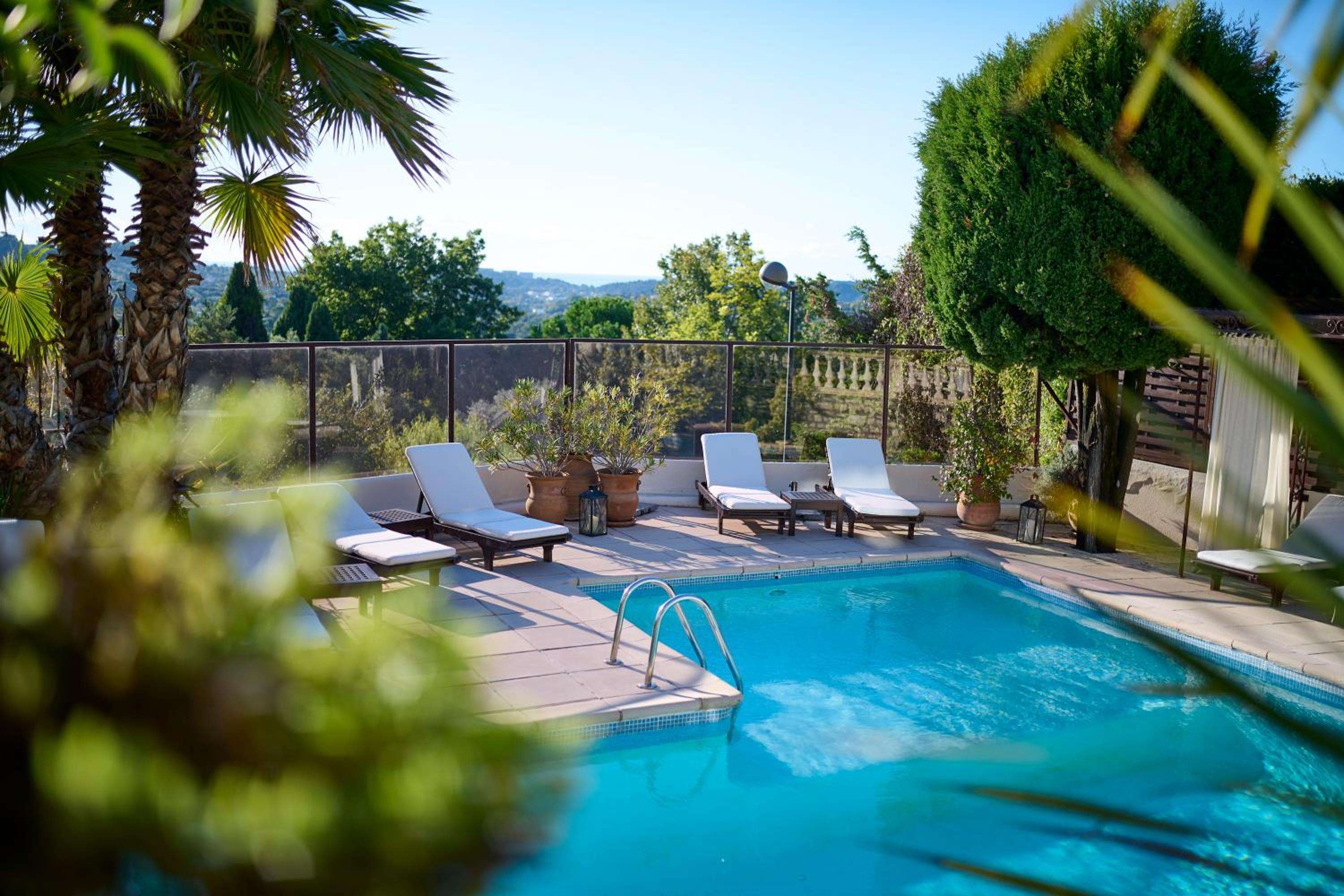 Sun-dappled loungers frame the azure pool where palm fronds cast dancing shadows, while terracotta pots and distant hills create an intimate Riviera sanctuary.