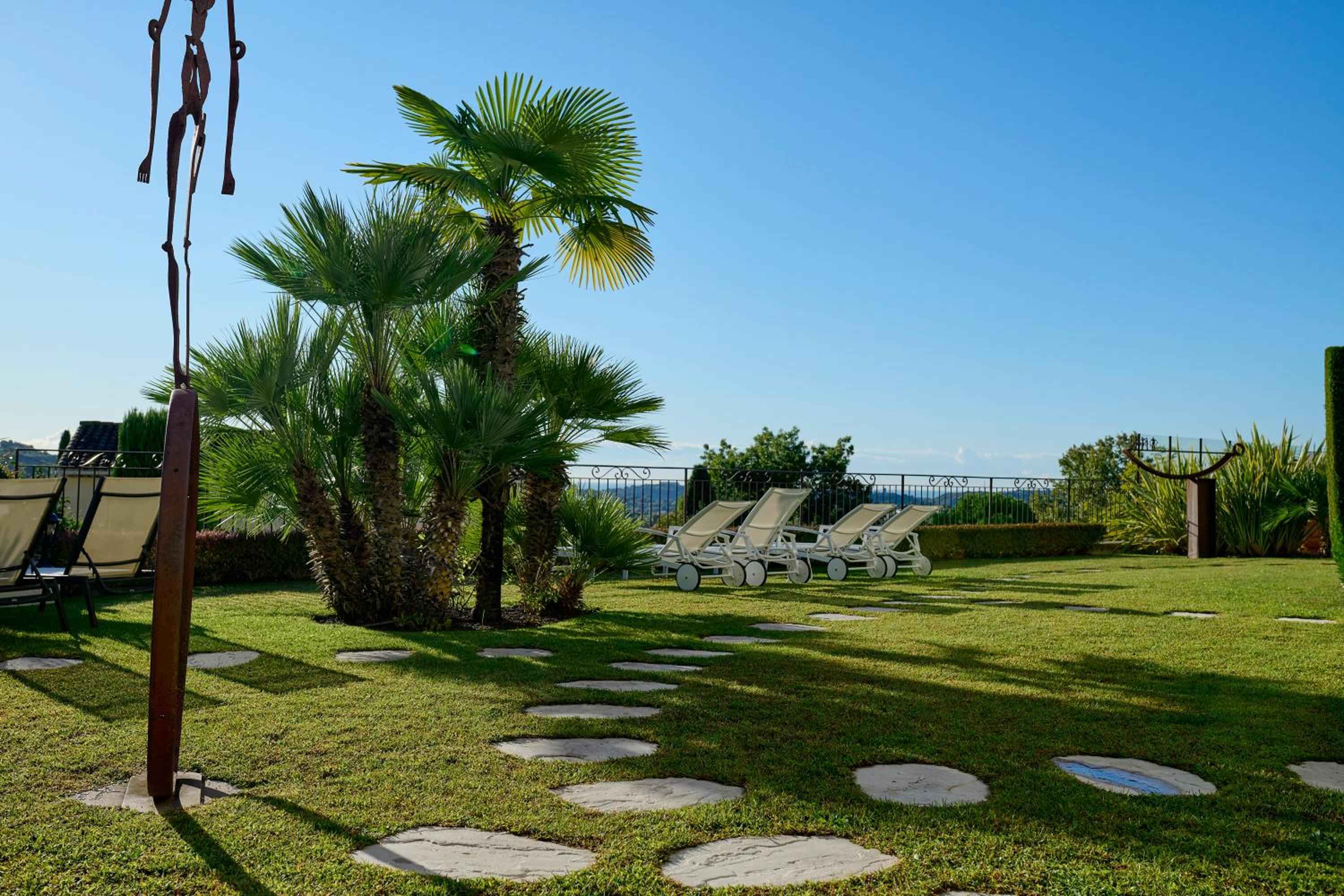 Stepping stones wind through manicured lawns toward sun loungers, where Mediterranean palms cast dappled shadows and iron railings frame distant azure views.