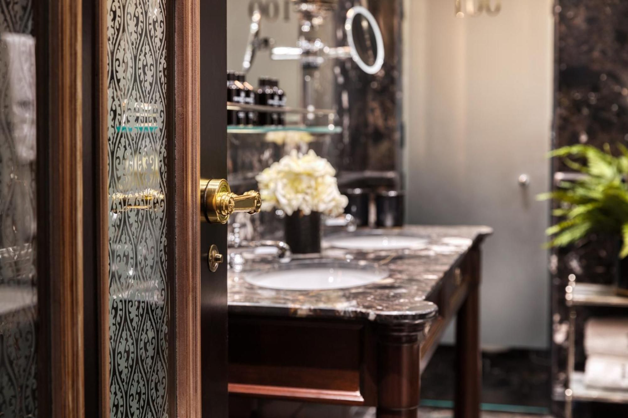 Brass fixtures gleam against etched glass doors, where marble vanities and fresh white blooms create an intimate sanctuary of refined luxury.