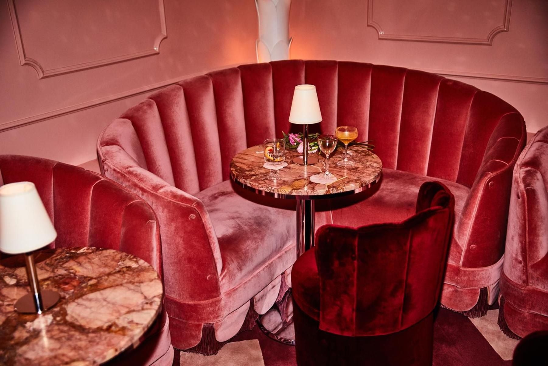 Plush rose velvet banquettes curve around marble-topped tables, creating intimate alcoves where champagne flutes catch the warm glow of delicate table lamps.