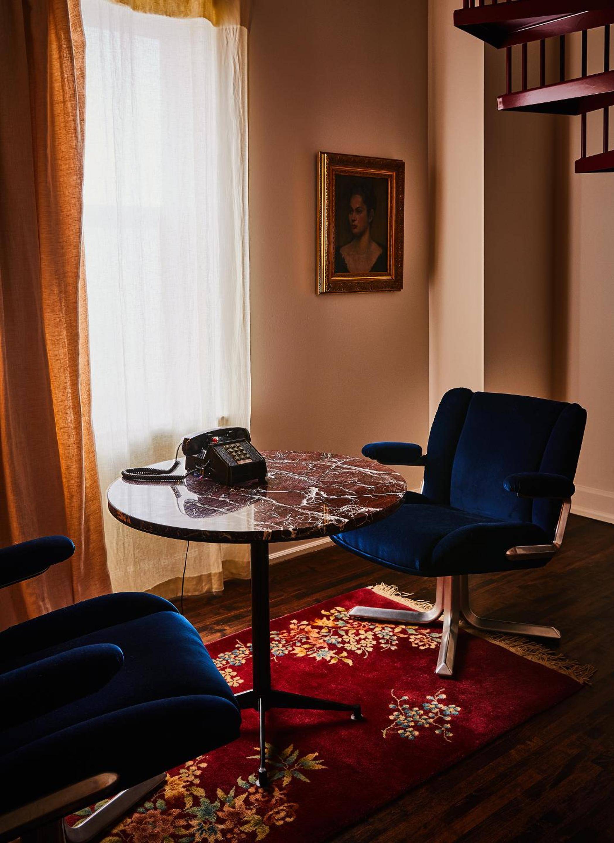 Navy velvet chairs and marble-topped table create an intimate corner where vintage rotary phone conversations unfold beneath golden afternoon light and classical portraiture.