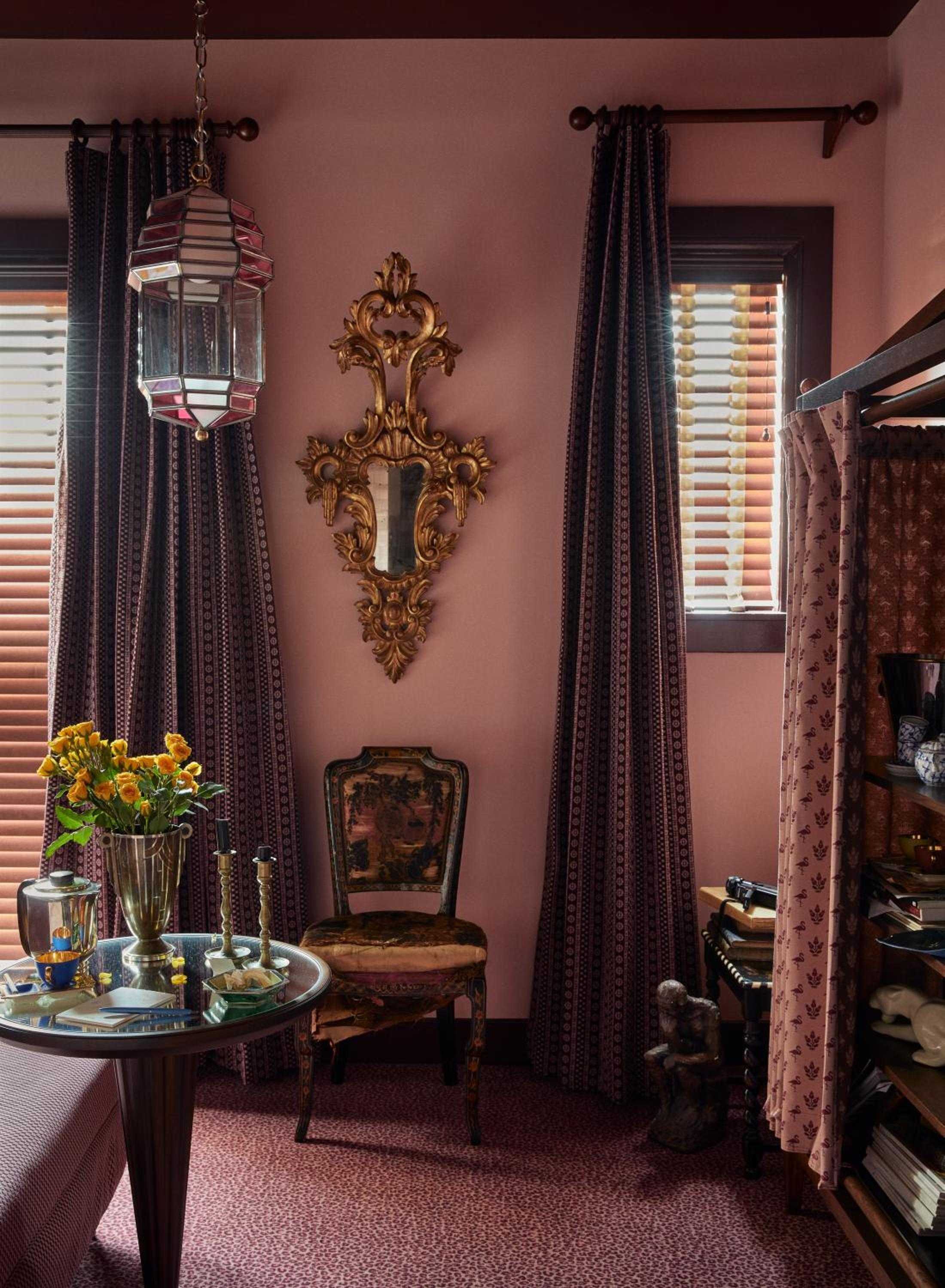 Dusty rose walls embrace an ornate gilded mirror and vintage chair, while fresh blooms catch morning light filtering through wooden blinds in this intimate corner.