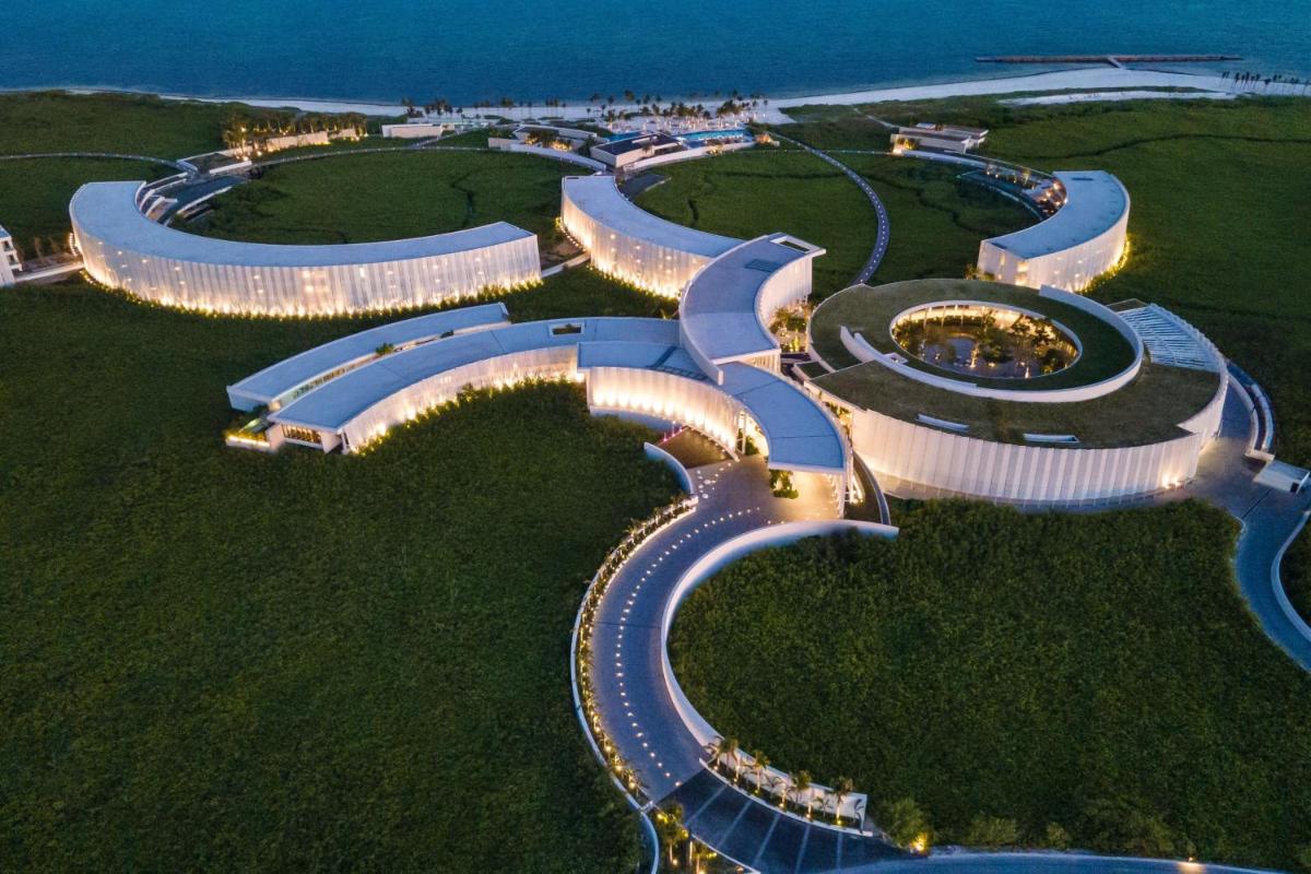 Serpentine white buildings flow like ocean waves across emerald jungle, their illuminated curves creating an otherworldly resort landscape against the Caribbean coastline at dusk.