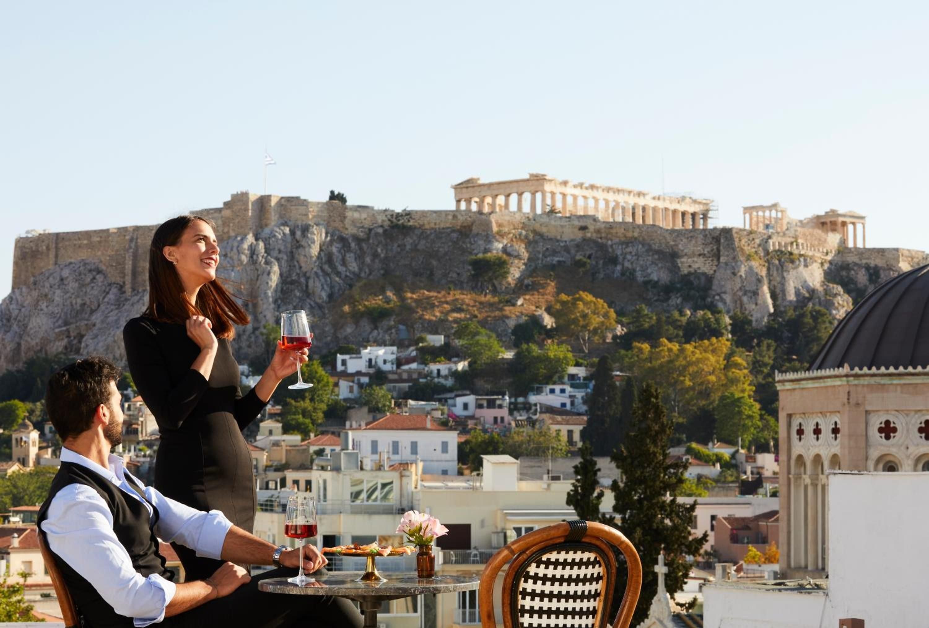 Sunset drinks unfold against Athens' crown jewel as couples toast golden hour from Villa Brown's terrace, the illuminated Acropolis creating an unforgettable backdrop.