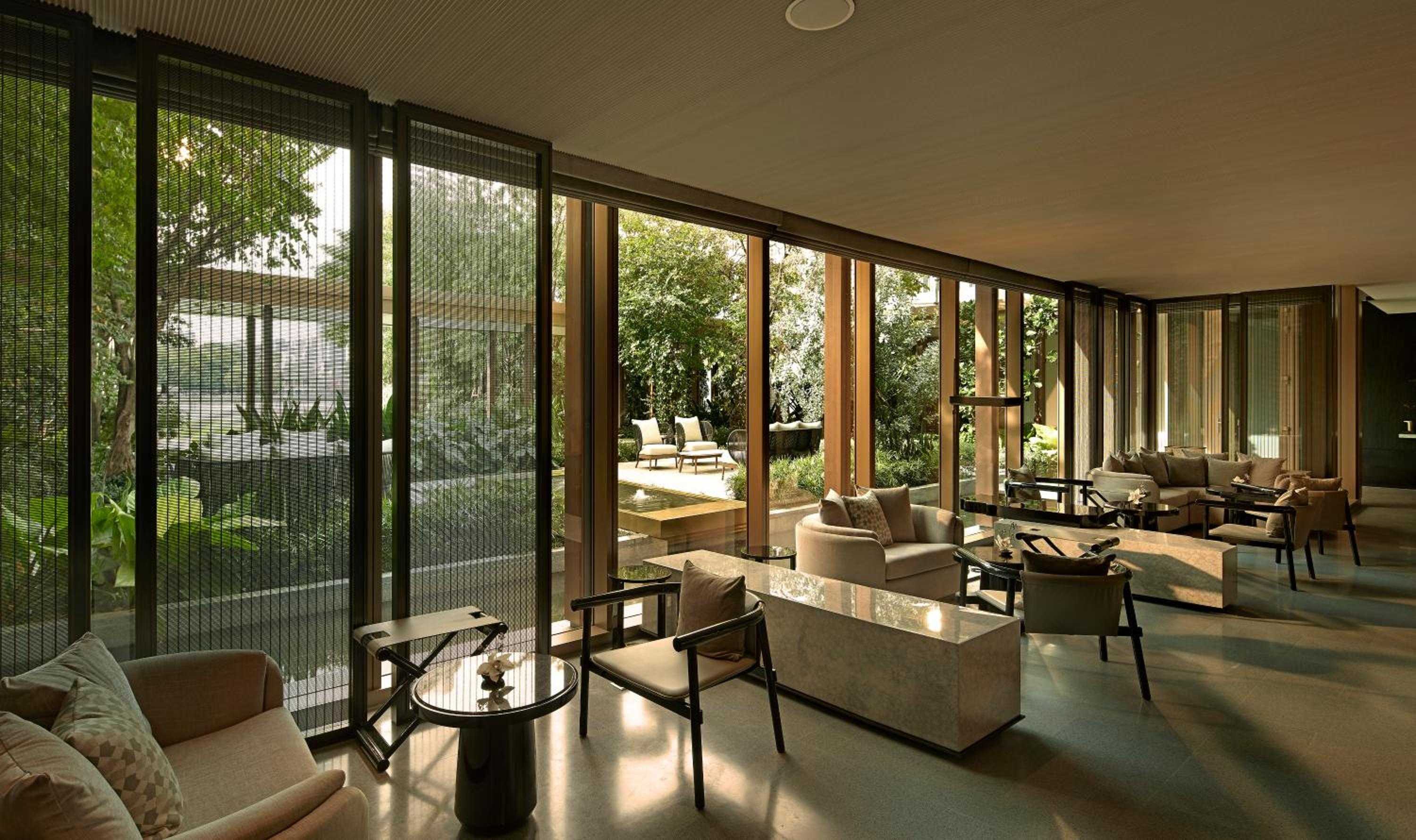 Floor-to-ceiling windows frame lush gardens while bronze mesh screens filter golden light across the sophisticated lounge's neutral palette and polished concrete floors.