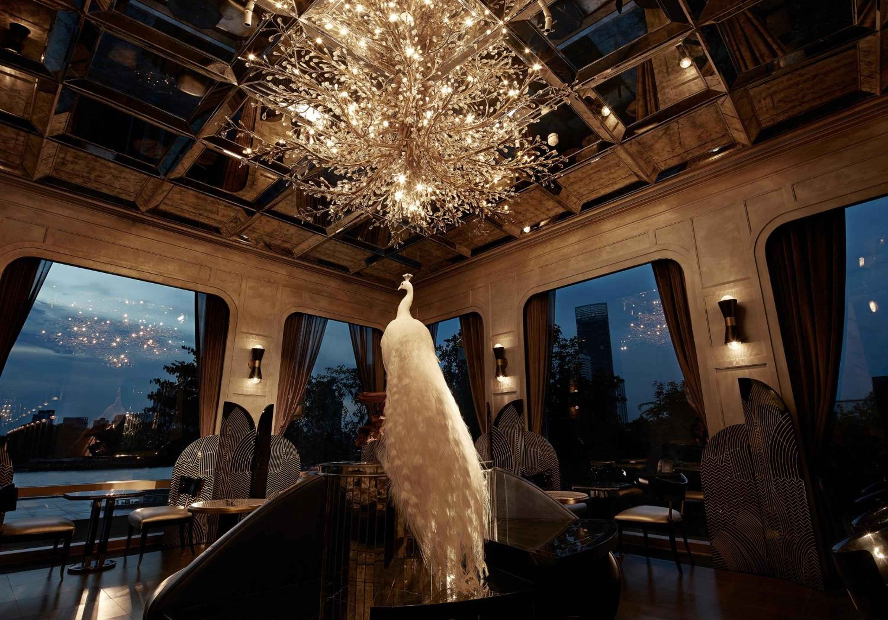 An albino peacock presides regally over Capella Bangkok's enchanting dining room, where intricate chandeliers cast golden light against panoramic Chao Phraya River views at twilight.