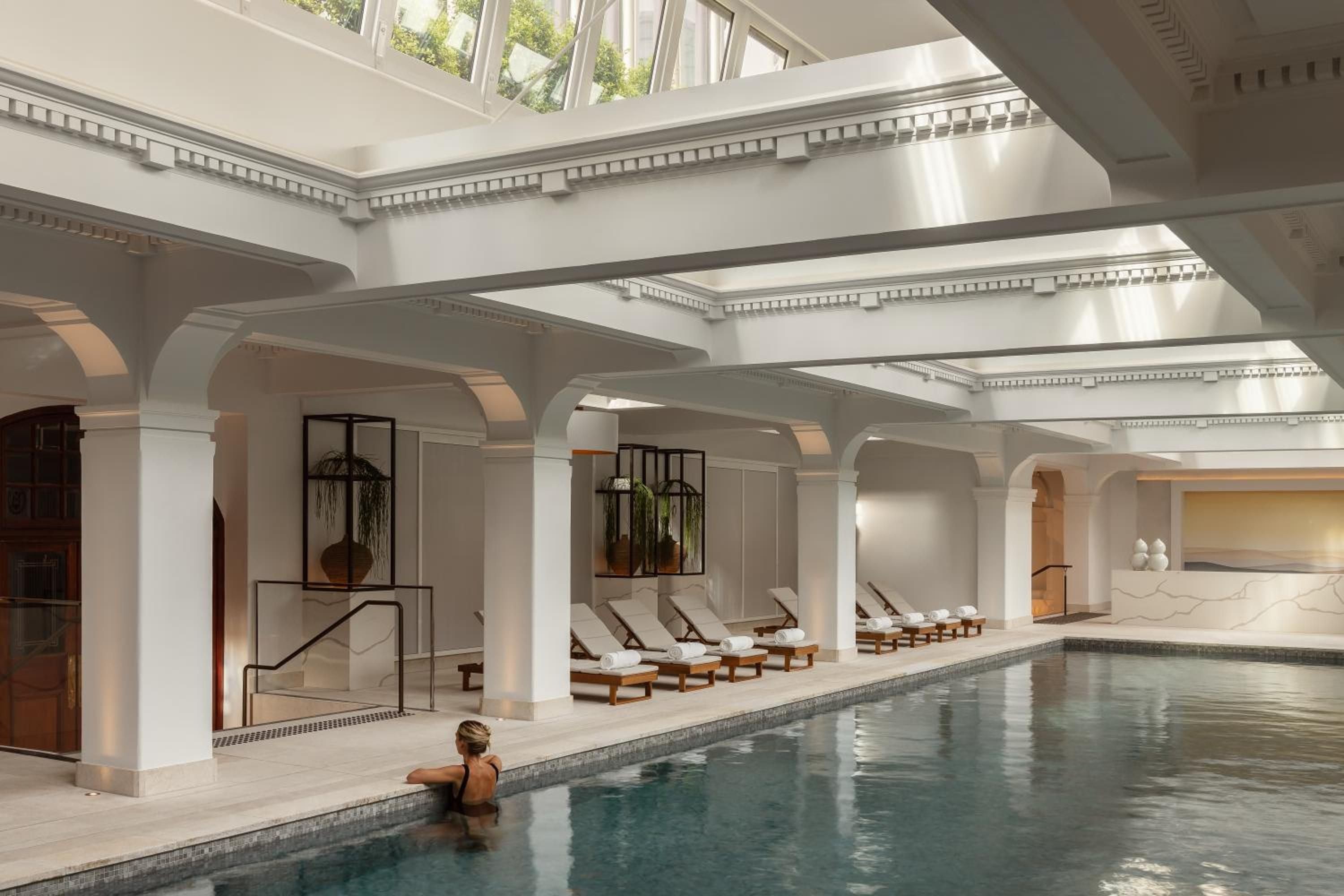 Classical columns frame the serene spa pool where natural light filters through clerestory windows, creating an elegant Roman bath atmosphere with marble details and poolside loungers.