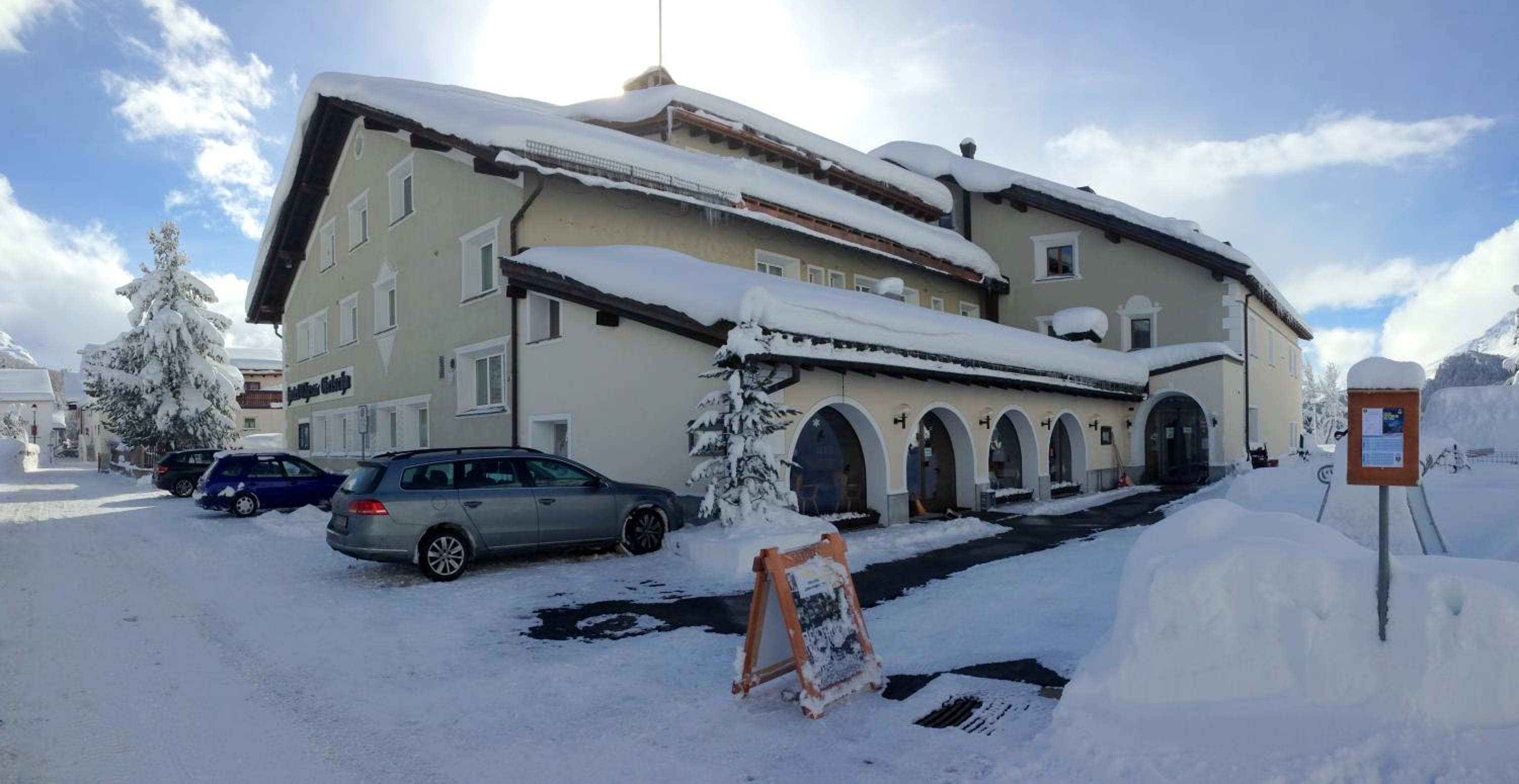 Snow blankets the charming alpine façade of Chesa Marchetta, where traditional arched entrances and winter-dusted eaves create an inviting mountain retreat against crisp blue skies.