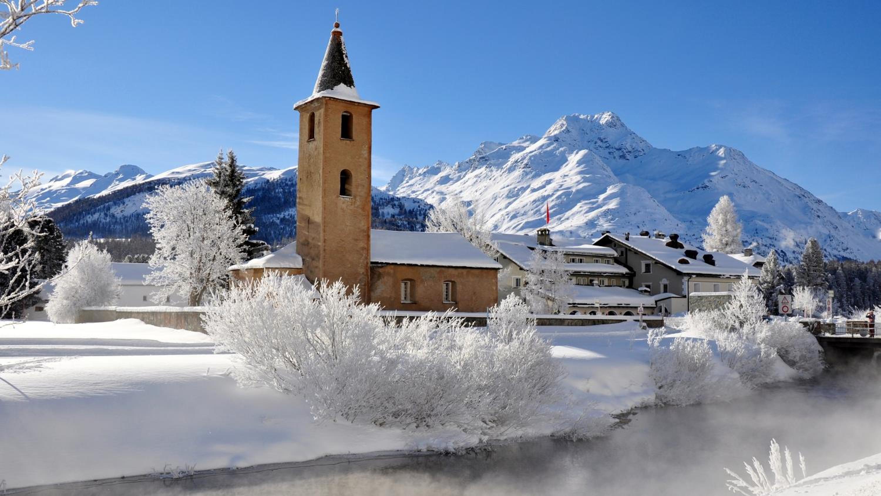 Frost-kissed village charm unfolds beneath towering Alpine peaks, where the historic bell tower stands sentinel over snow-blanketed rooftops and steaming winter air.
