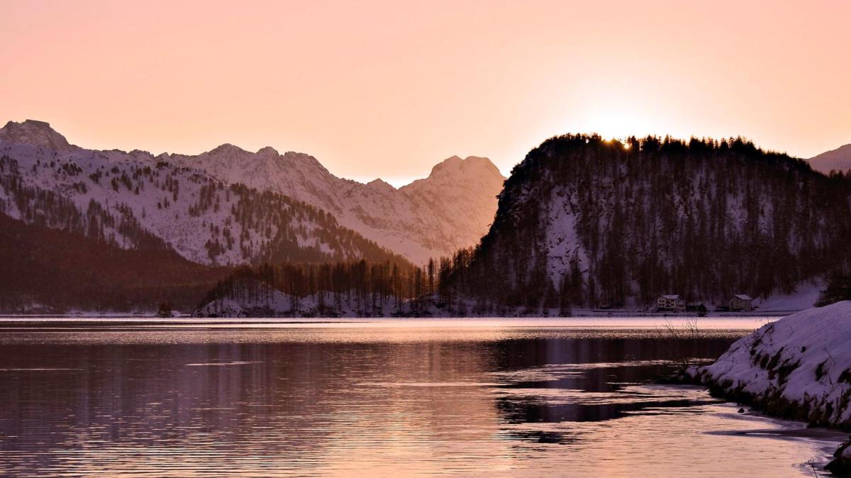 Alpine peaks catch dawn's rosy glow above mirror-still waters, where snow-dusted shores and gentle ripples create a scene of profound mountain tranquility.