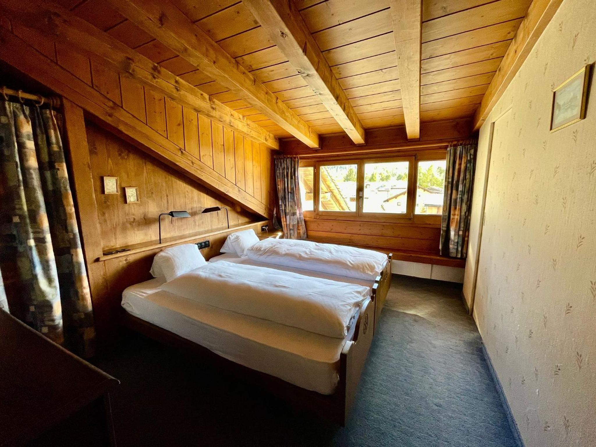 Exposed timber beams and weathered wood walls create an intimate alpine retreat, where twin beds nestle beneath slanted eaves overlooking rooftops through curtained windows.