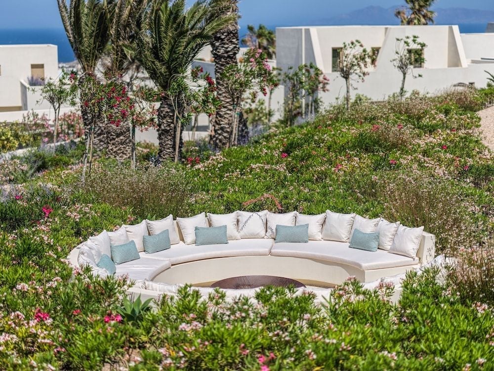 Sunken seating carved into volcanic stone creates an intimate amphitheater amid native Mediterranean flora, where turquoise cushions echo the Aegean beyond white-washed villas.