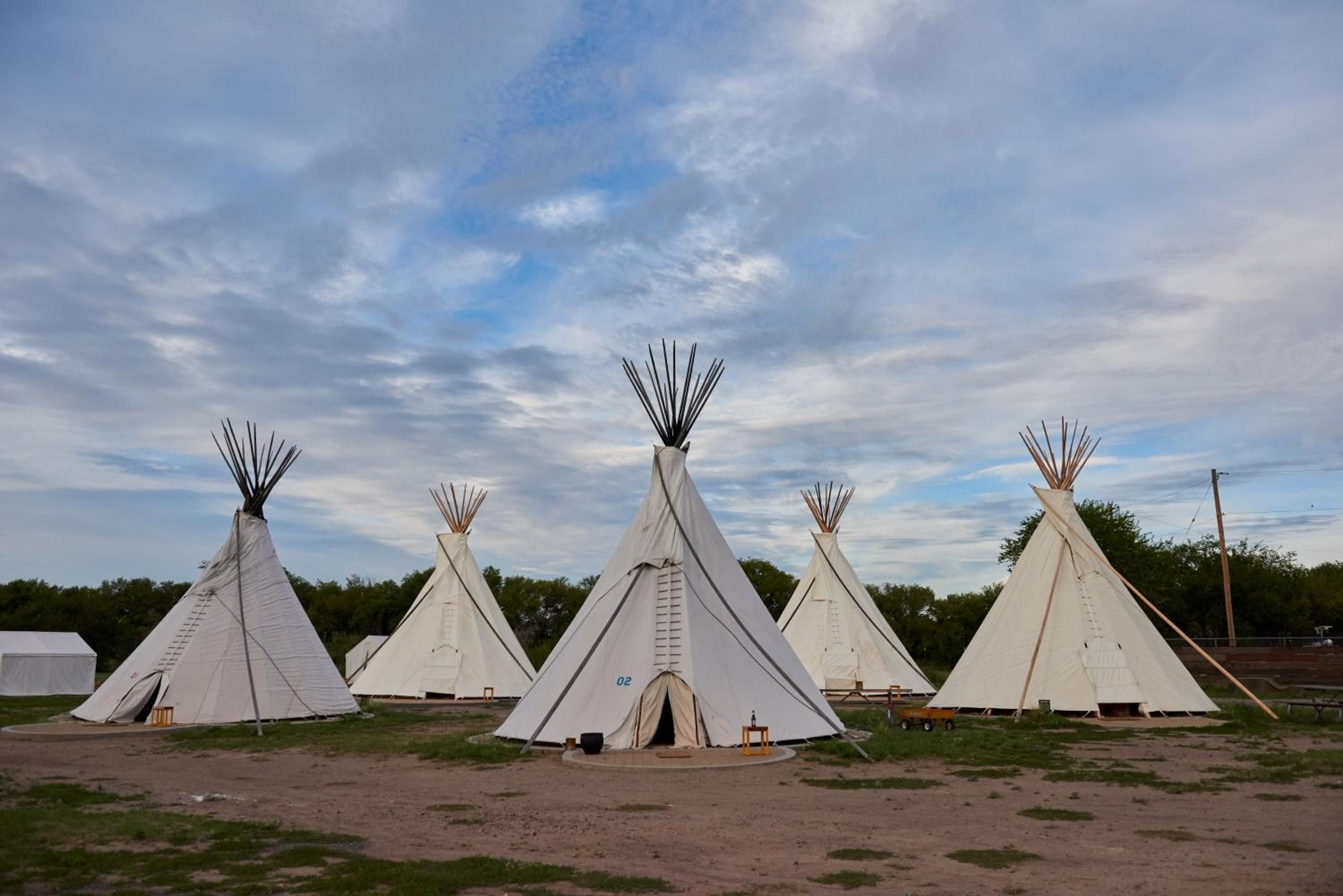 Canvas tepees dot the high desert landscape under vast Texas skies, where traditional lodge poles pierce cloudy horizons and glamping meets authentic frontier spirit.