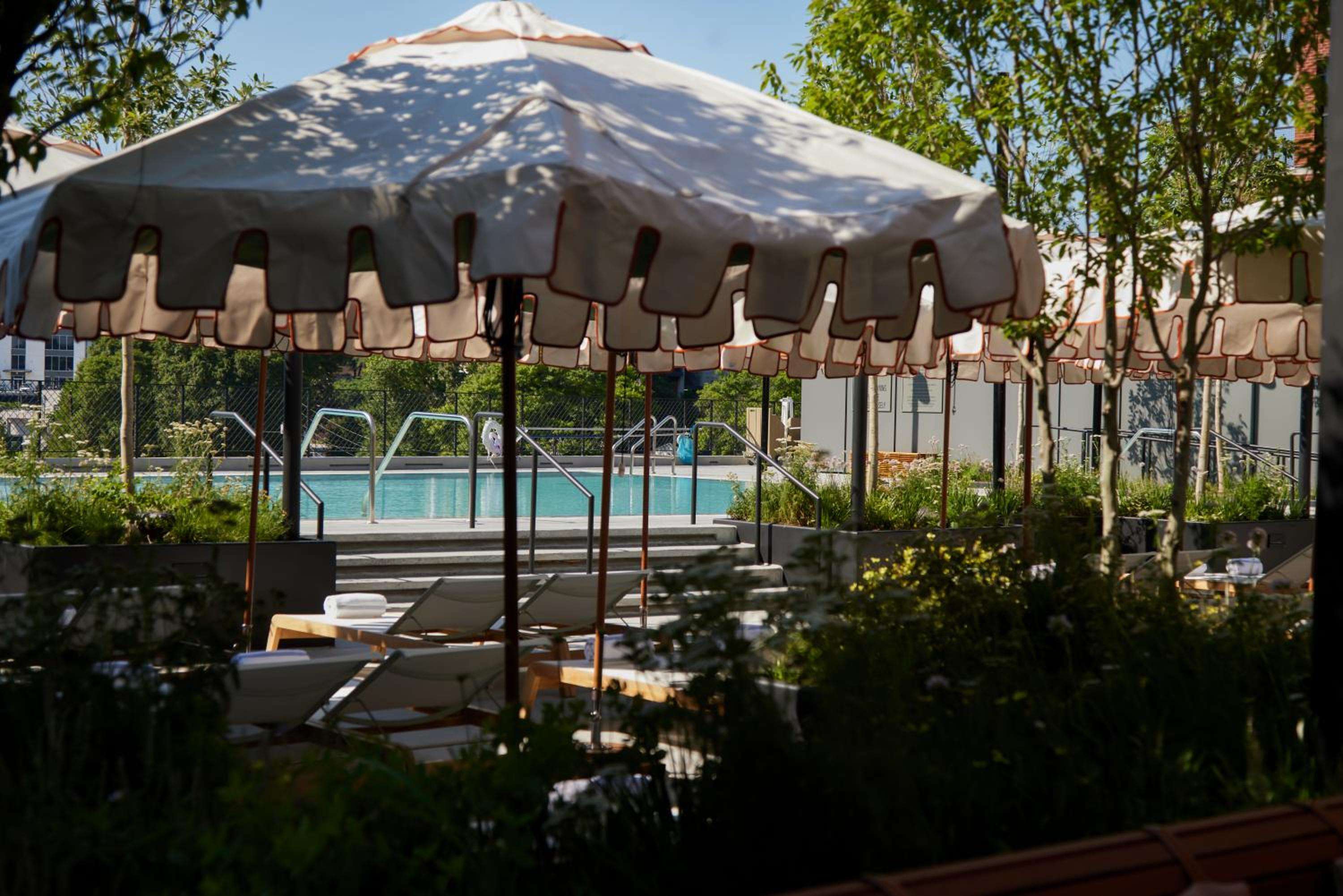 Dappled sunlight filters through leafy canopy onto cream-striped umbrellas, where poolside loungers beckon beneath swaying branches in this tranquil urban oasis.