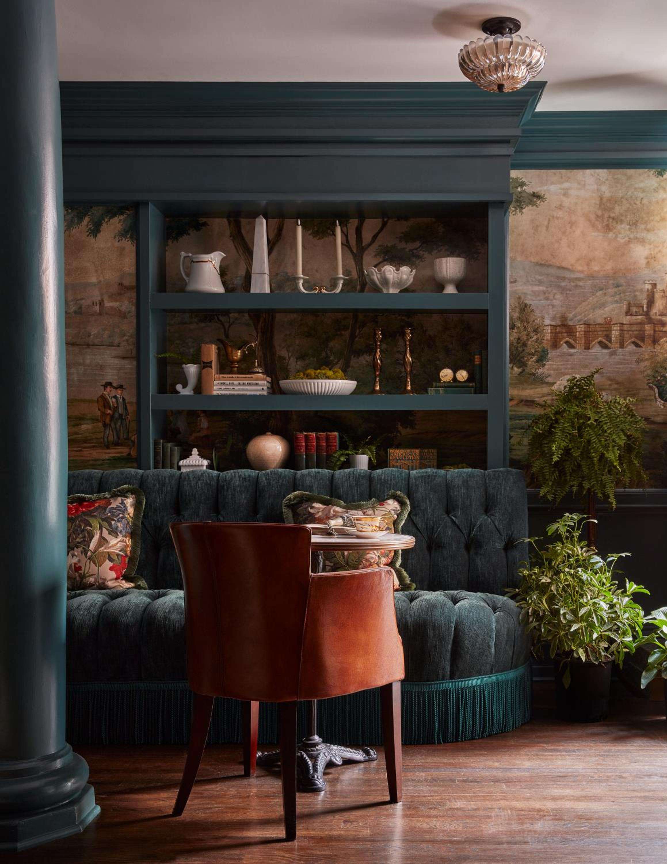 Emerald built-ins frame a tufted banquette where guests settle into cognac leather chairs, surrounded by pastoral murals and carefully curated ceramics in this intimate corner.