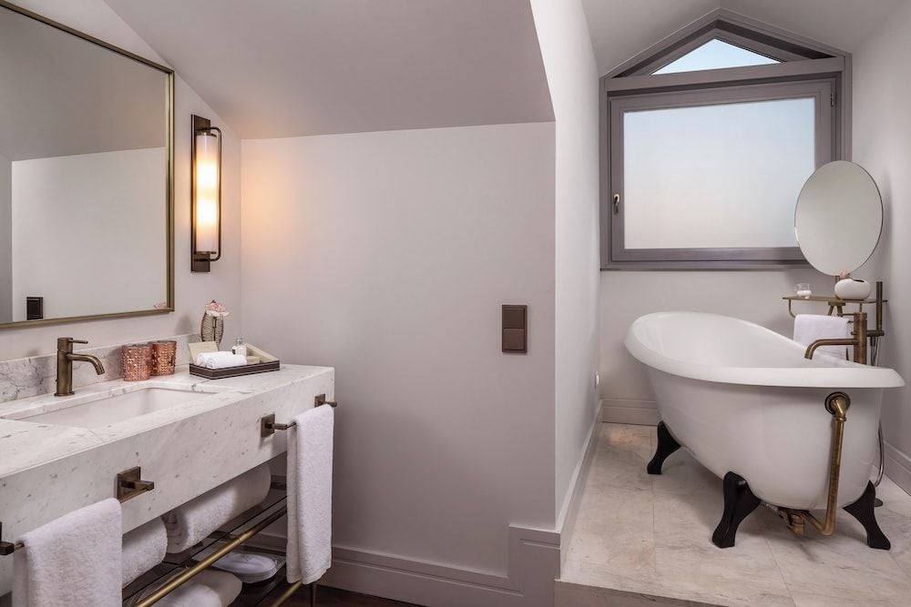 Marble surfaces gleam under warm sconce lighting while a sculptural clawfoot tub anchors this serene attic bathroom, where slanted ceilings create intimate luxury.