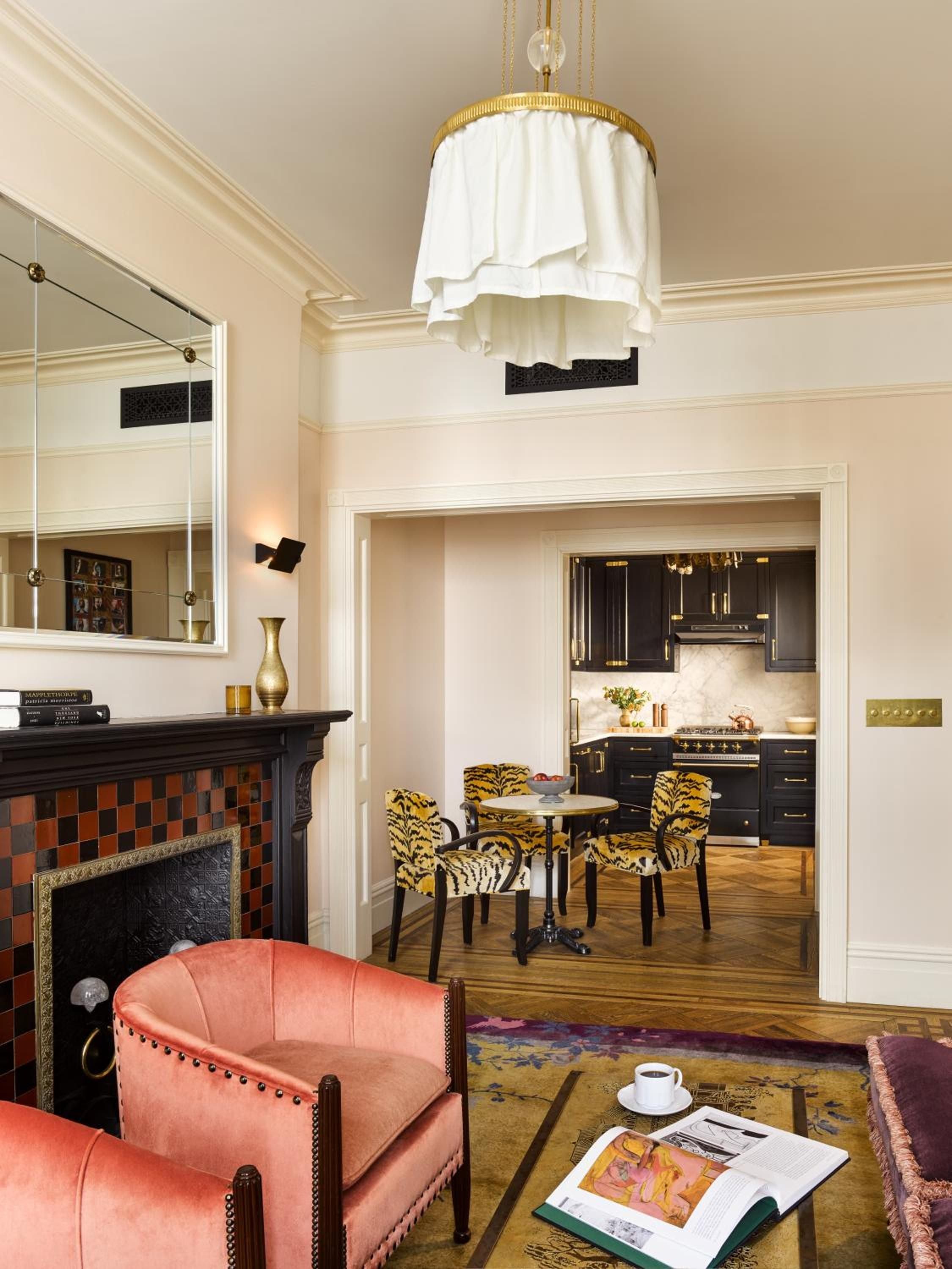 Crown molding frames a sophisticated living space where coral leather meets tiger-print dining chairs, while soft pendant lighting illuminates the passage to a sleek galley kitchen.