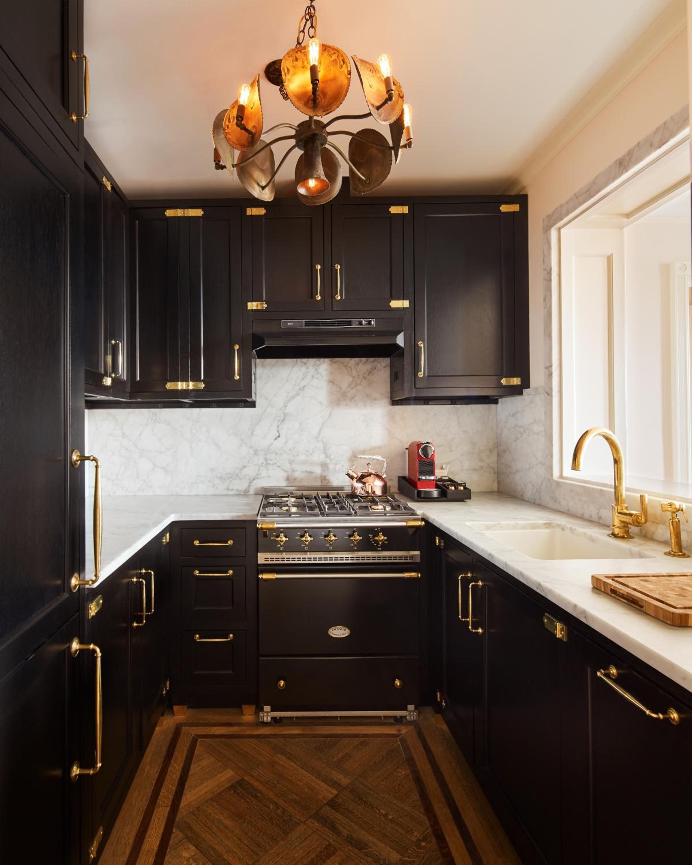 Moody black cabinetry and brass fixtures create intimate elegance in this compact galley kitchen, where marble backsplash catches golden light from the sculptural chandelier above.