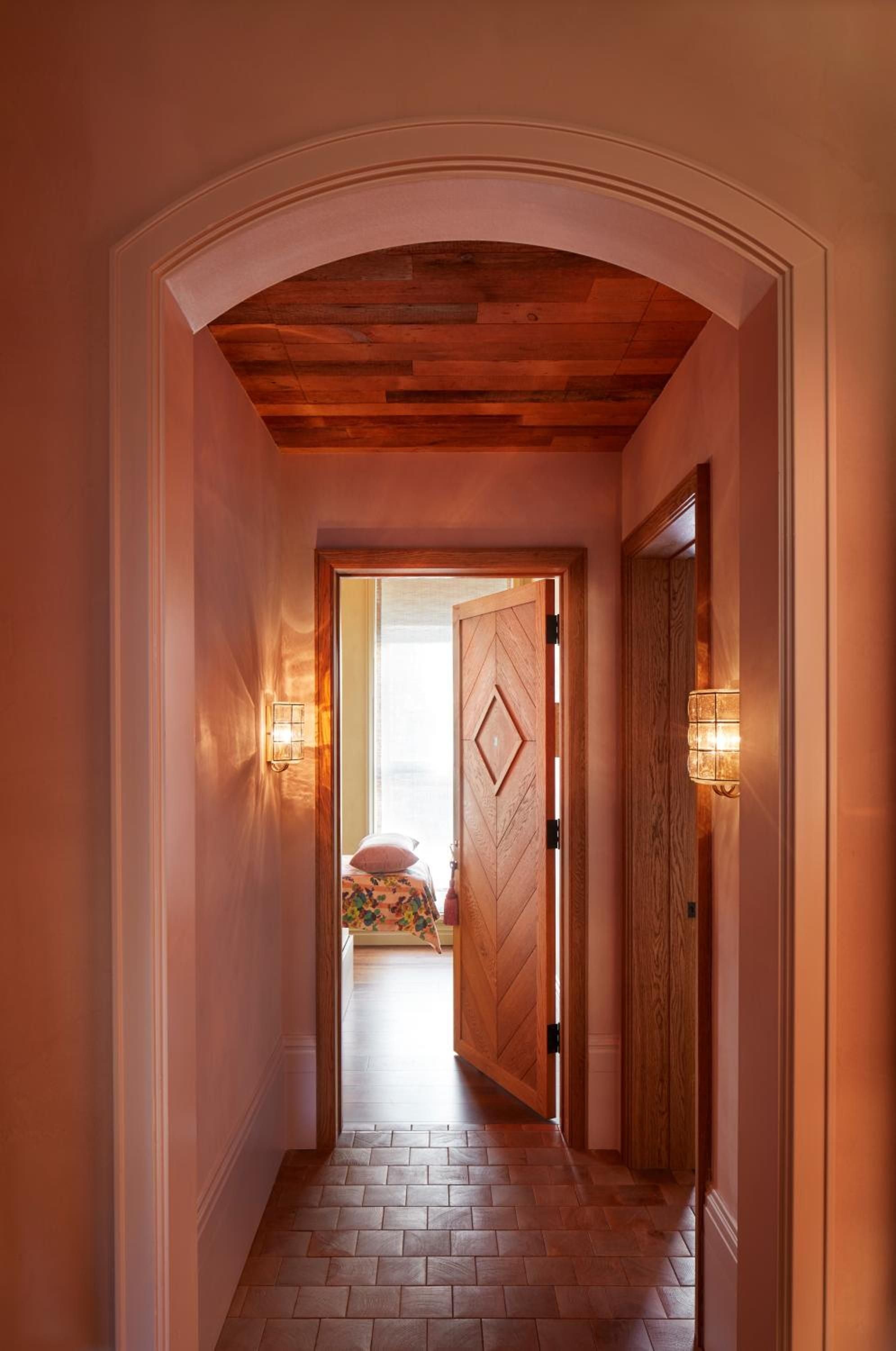 Warm terracotta walls and exposed wooden beams frame an intimate passageway, where geometric door patterns and glowing sconces create an inviting residential ambiance.