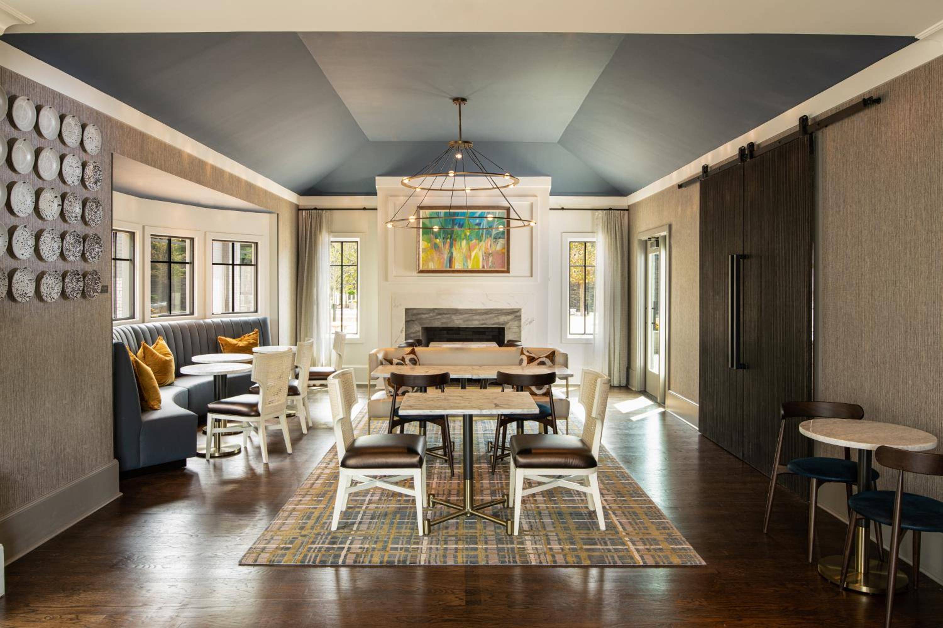 Natural light floods the contemporary dining space where plaid rugs anchor intimate seating arrangements beneath soaring coffered ceilings and geometric chandeliers.