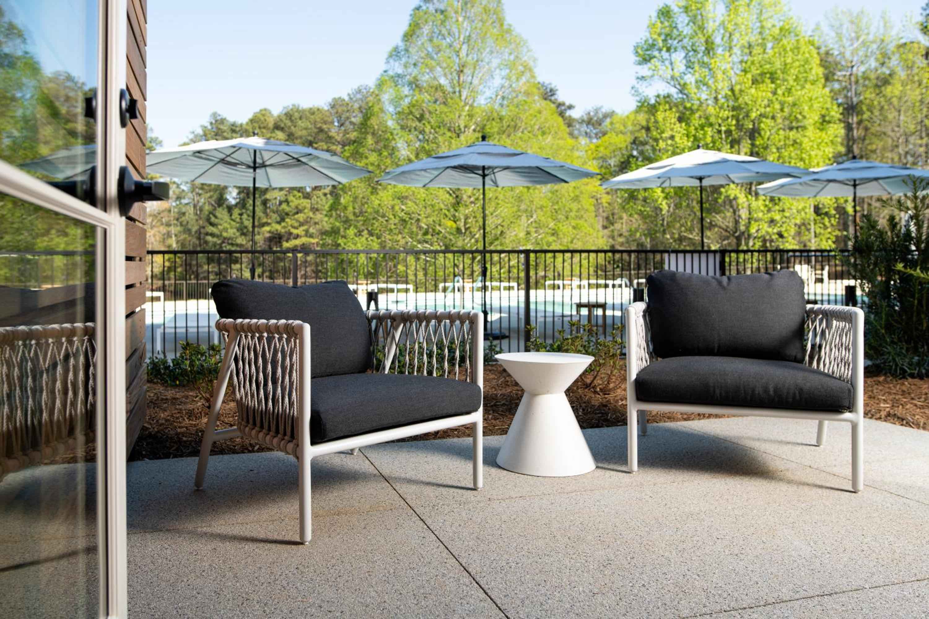 Contemporary rope-detailed chairs overlook the sparkling pool area, where umbrella shade dances across manicured landscaping and modern outdoor lounging beckons beneath towering trees.
