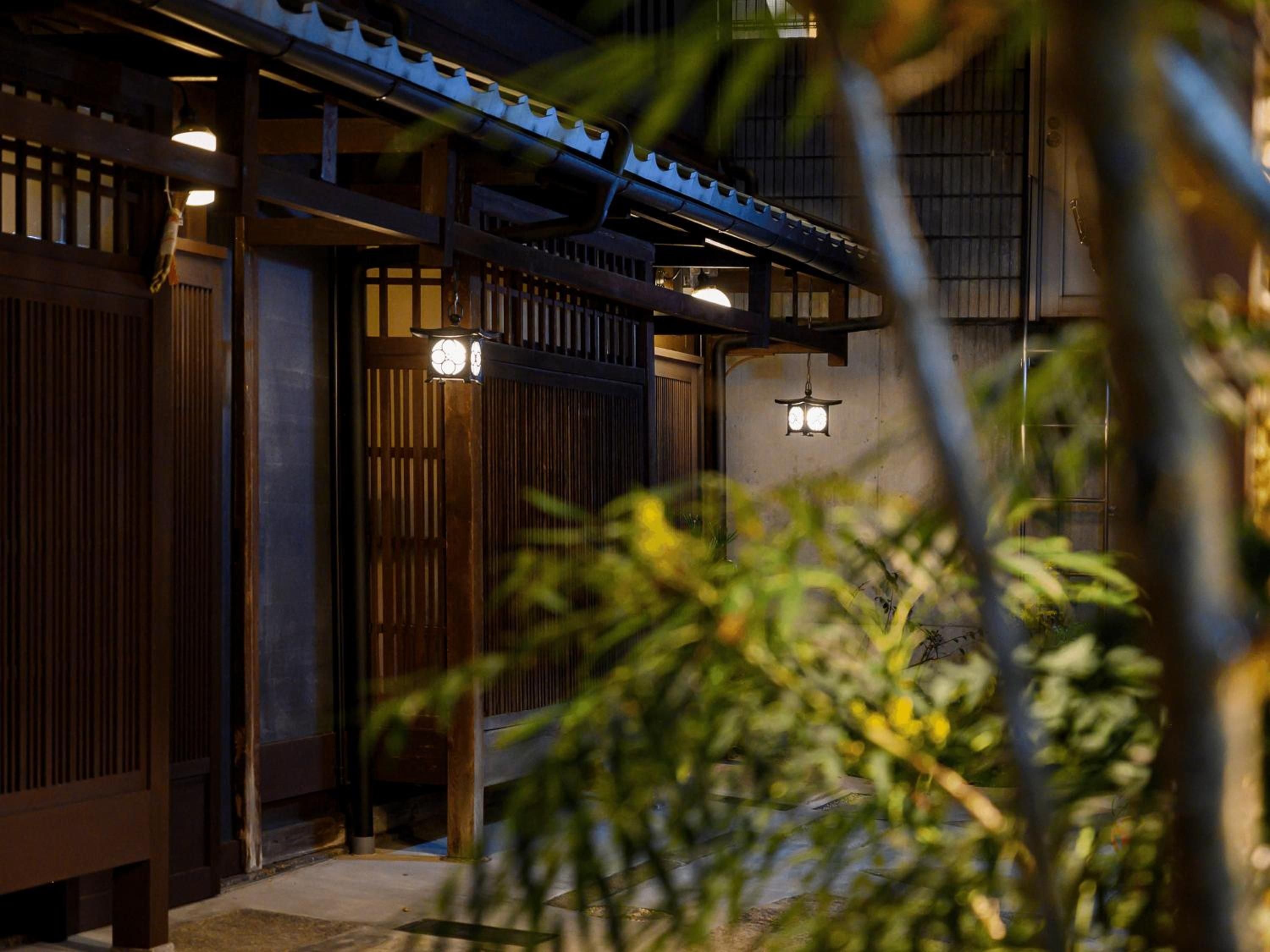 Traditional wooden lattice screens and glowing lanterns create an intimate evening ambiance along the machiya's authentic Kyoto streetscape, where centuries-old craftsmanship meets modern hospitality.
