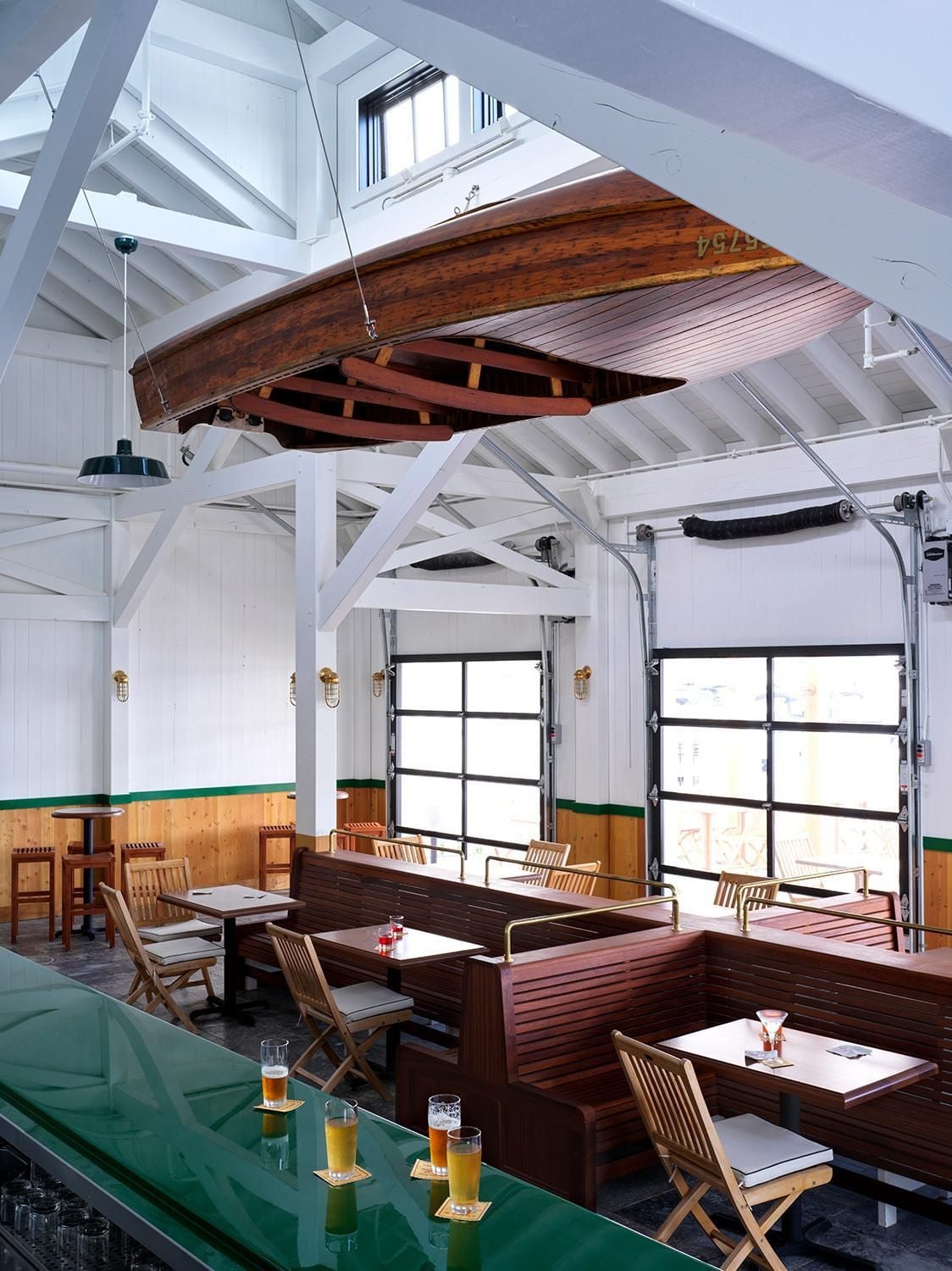 Suspended vintage boat and garage doors create nautical charm in this waterfront dining room, where craft beer flows beneath exposed white beams and warm wood accents.