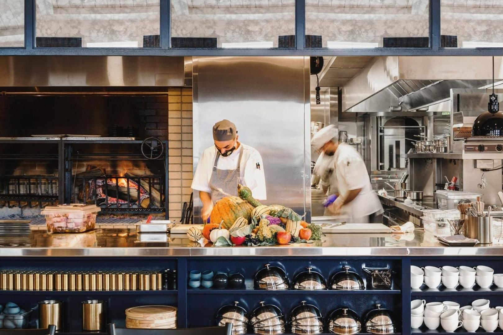 Chefs work their craft behind gleaming steel counters while seasonal bounty awaits transformation in this theatrical open kitchen where flames dance and culinary artistry unfolds.
