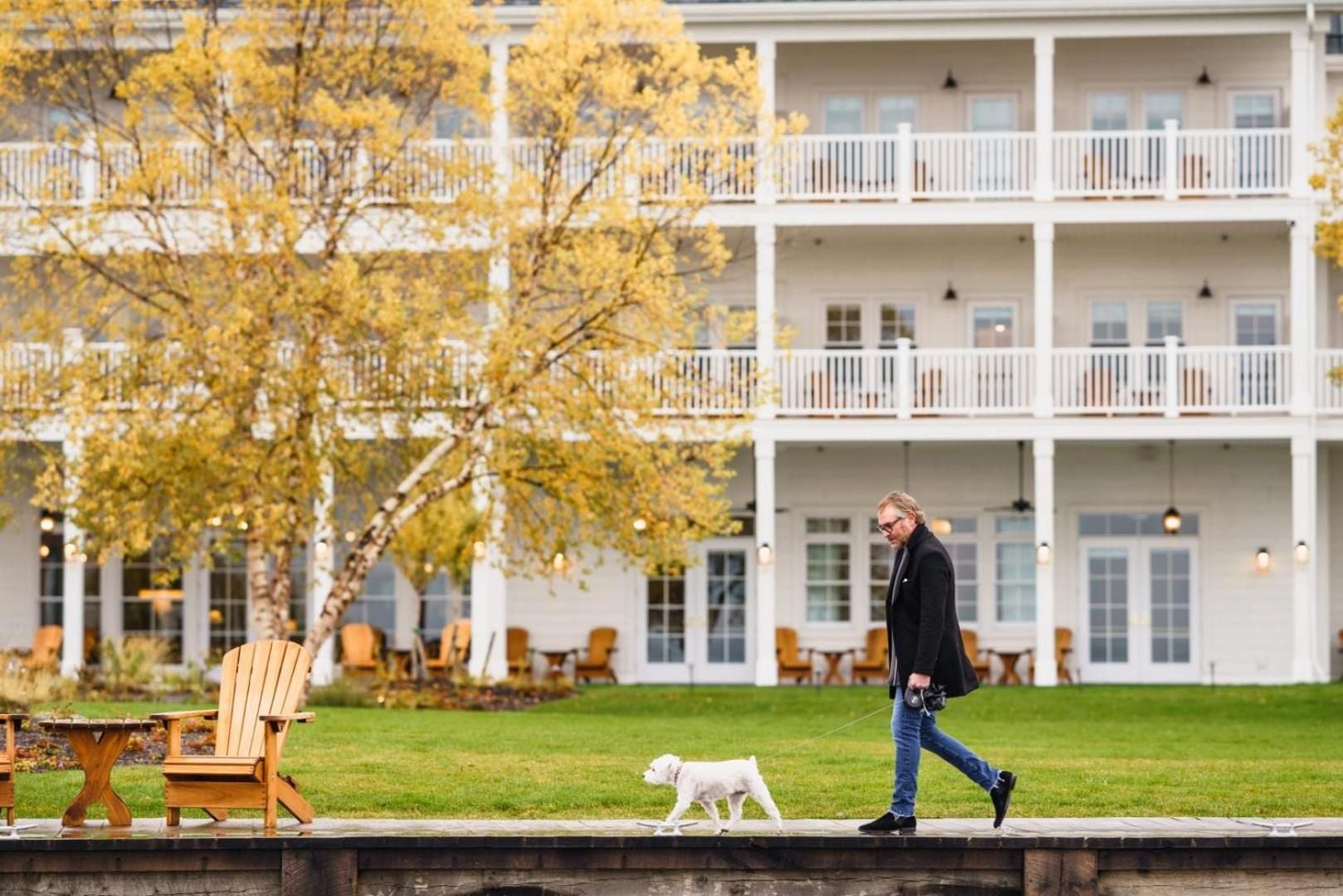 Golden autumn light filters through birch trees as guests stroll manicured lawns with their companions, the resort's stately white balconies creating an elegant New England backdrop.