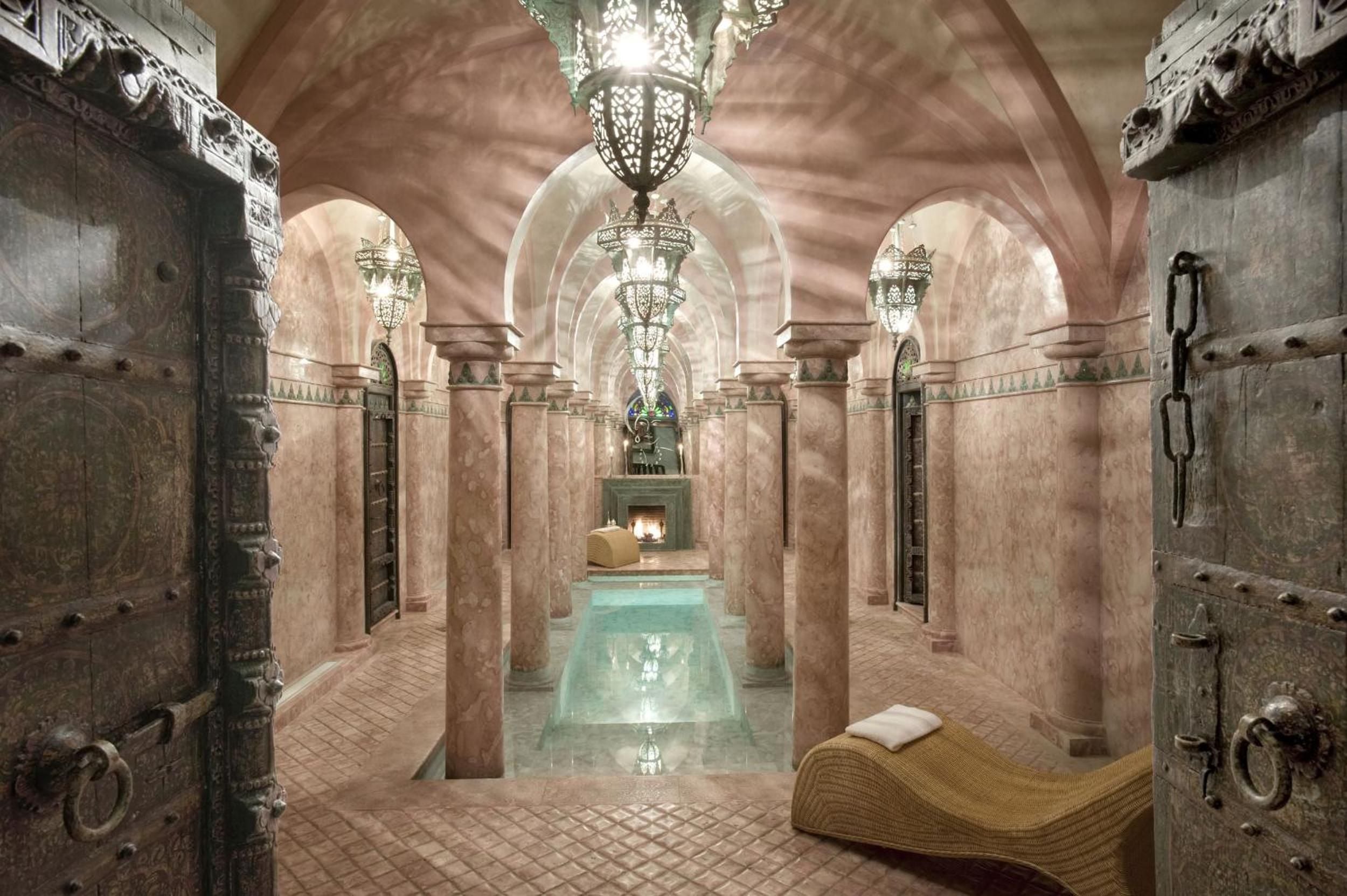 Moorish arches frame a serene plunge pool in the hammam, where ornate lanterns cast intricate shadows across warm stone columns and traditional tilework.