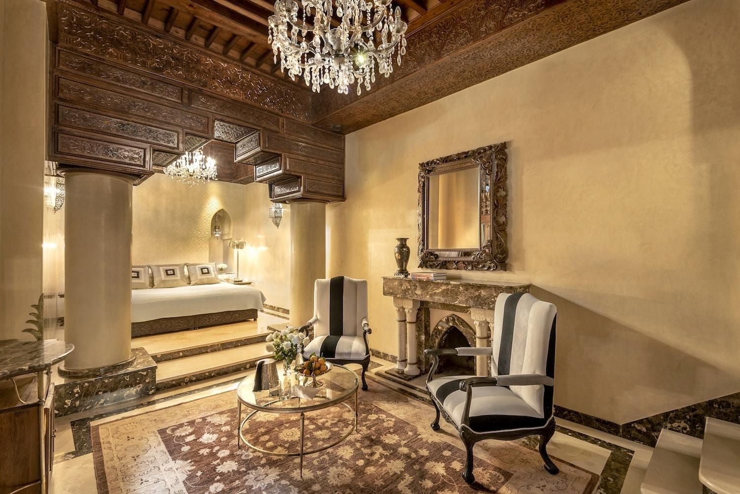 Intricate carved cedar arches frame this palatial suite, where crystal chandeliers cast warm light across hand-painted ceilings and striped silk chairs flanking an ornate marble fireplace.