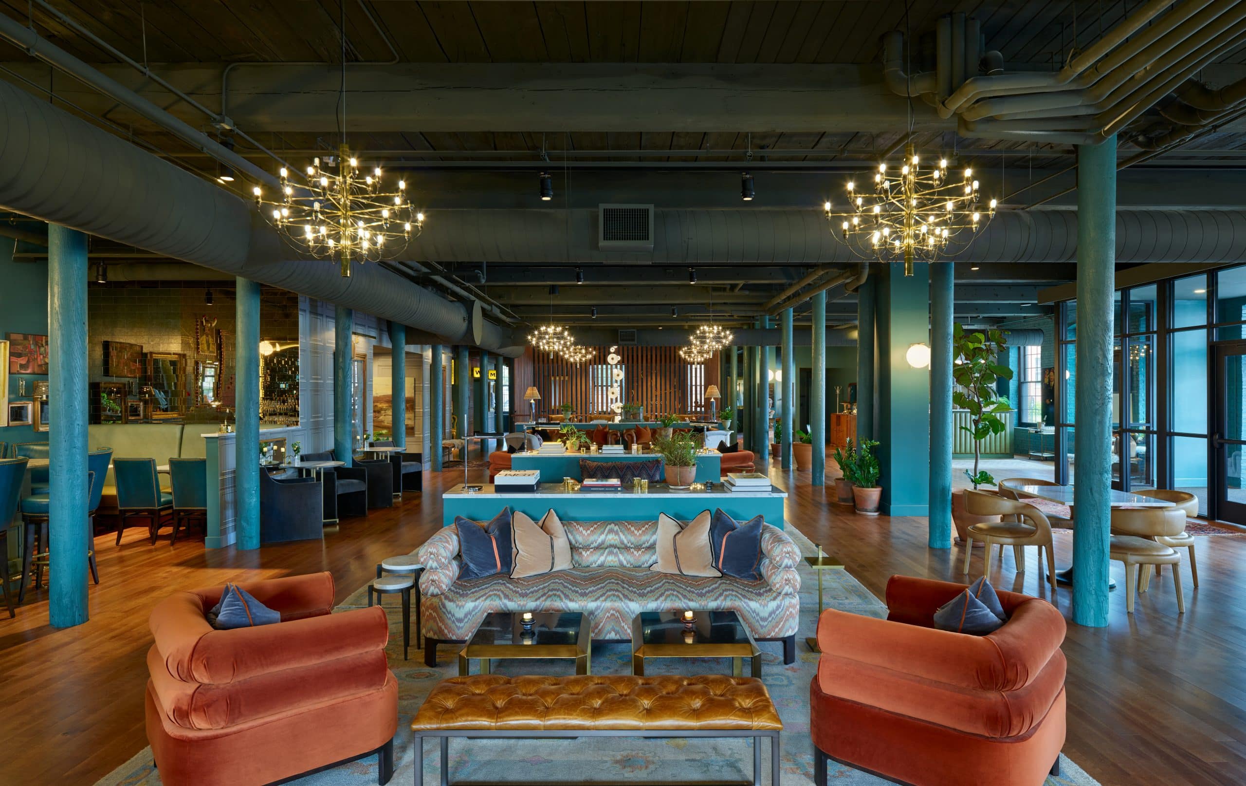 Industrial chic meets residential comfort in this loft-like lobby, where teal columns frame conversation clusters beneath sculptural brass chandeliers and exposed ductwork.