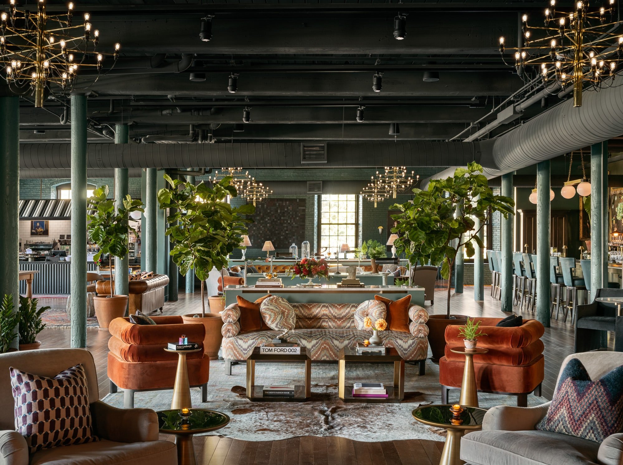 Burnt orange velvet chairs and geometric patterns create intimate conversation nooks beneath soaring industrial ceilings, while fiddle leaf figs add organic warmth to exposed brick walls.